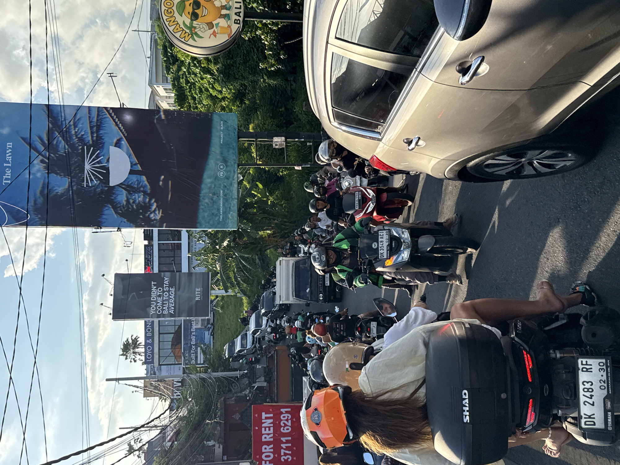 The image shows heavy traffic in what appears to be North Kuta, Indonesia. The view is from the back of a motorcycle with a passenger. The motorcycle's license plate is DK 2483 RF. Other vehicles, predominantly motorcycles, are visible in the packed street. Various signs and billboards flank the road, advertising "The Lawn", "Mango Bali", and rental properties. One billboard reads, "YOU DIDN'T COME TO BALI TO STAY AVERAGE." Overhead power lines are evident against a backdrop of blue sky and clouds.