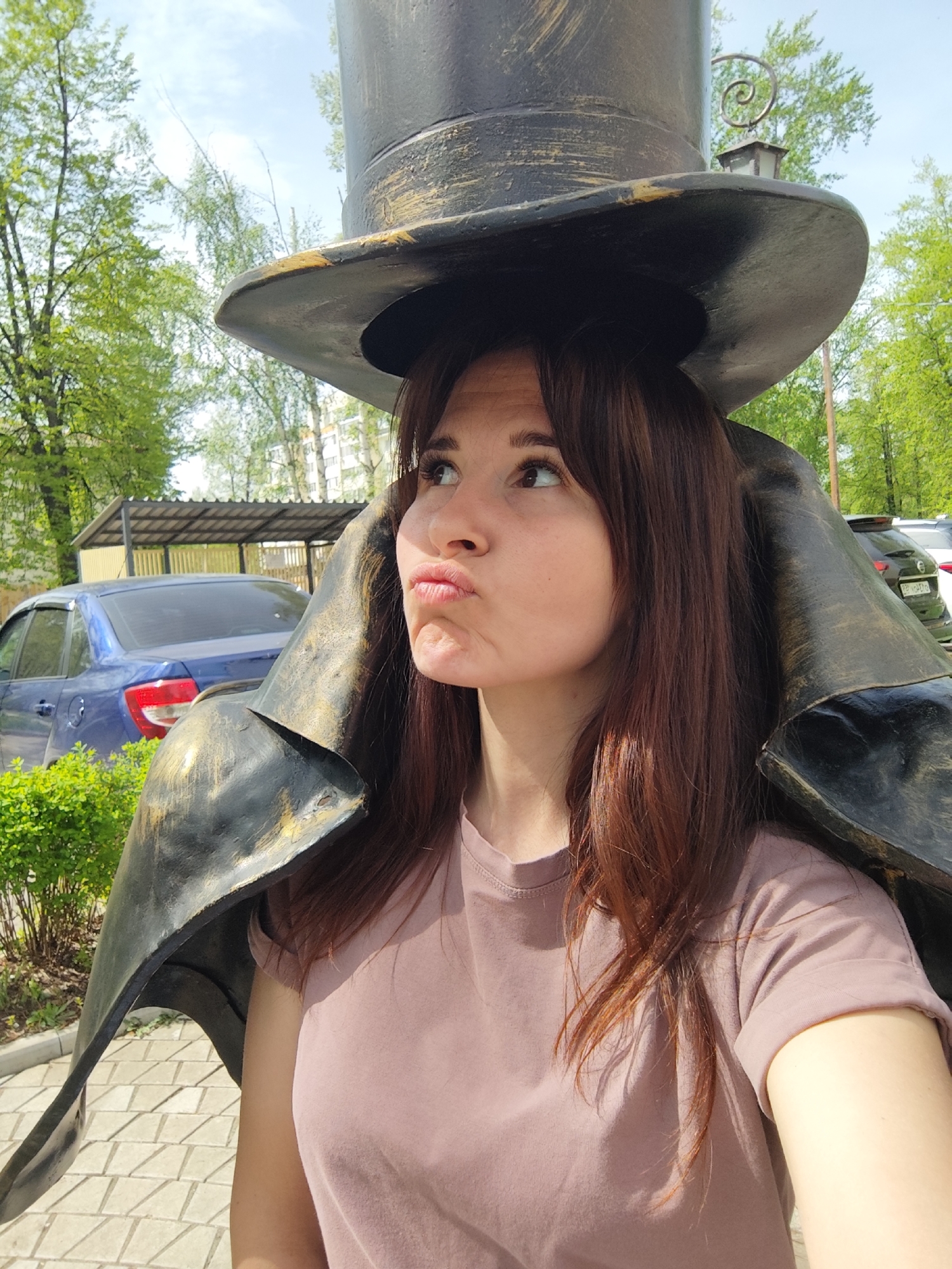 A young, light-skinned woman with brown hair is standing close to a statue in the city of Chaikovsky, Russia. The statue is that of a figure wearing a large black top hat and cape. The woman's head is positioned underneath the hat of the statue, creating the illusion that she is wearing it. She is wearing a pink t-shirt and has a playful expression on her face, with her lips pursed. The background shows a paved area, trees with green leaves, a blue car, and buildings. The photo appears to be taken during the daytime in a park or public area.