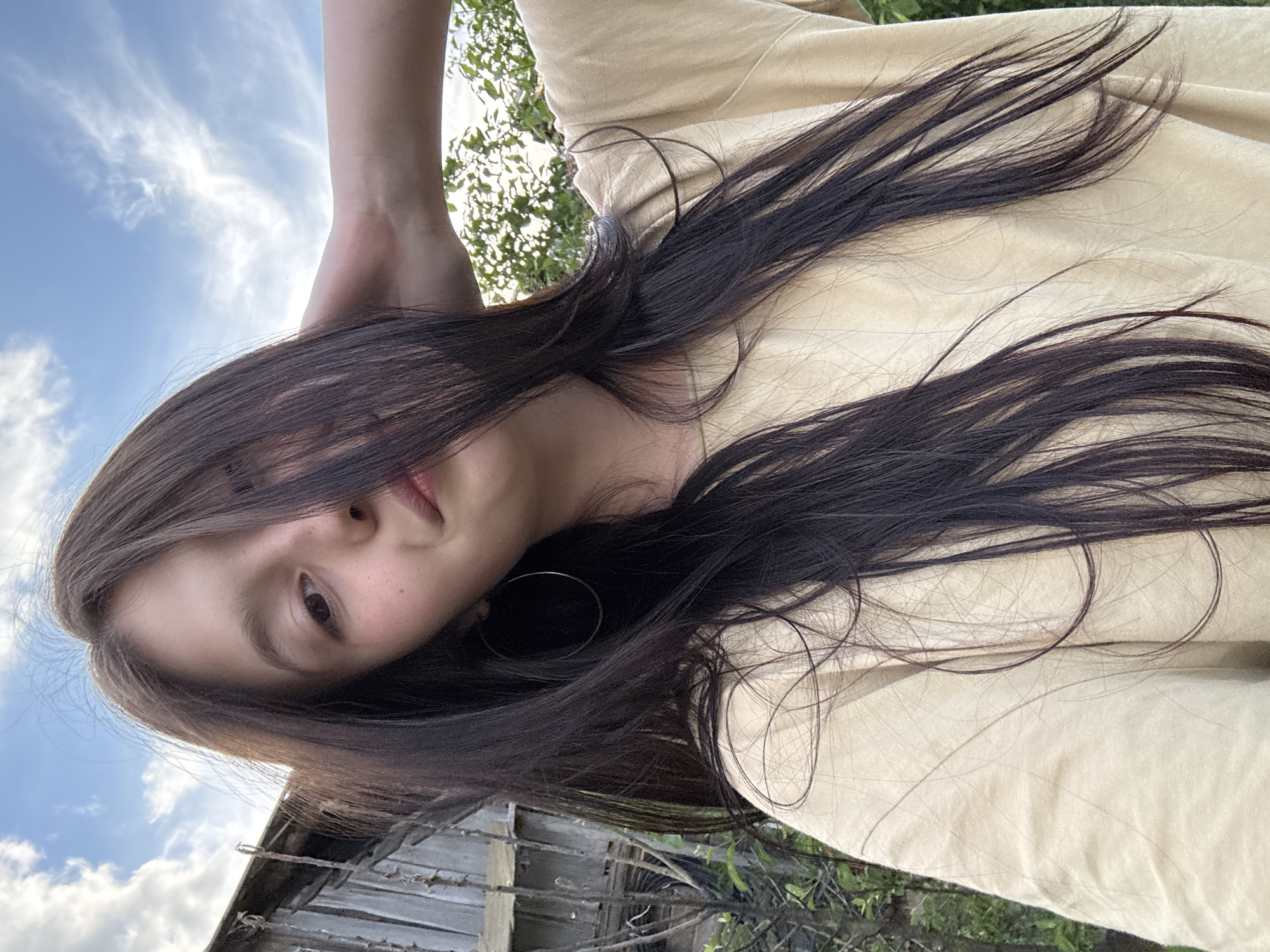 A person, presumably female, is photographed in an unusual perspective, likely from a lower angle looking upwards. The person has dark hair that partially obscures their face. They are wearing a light-colored shirt. An earring is visible. The background includes a patch of sky with clouds, some trees and foliage, and a weathered wooden structure, possibly a fence or shed. While the location is Фоки, Russia, the image does not provide specific urban elements to detail beyond the general rural/outdoor impression conveyed.