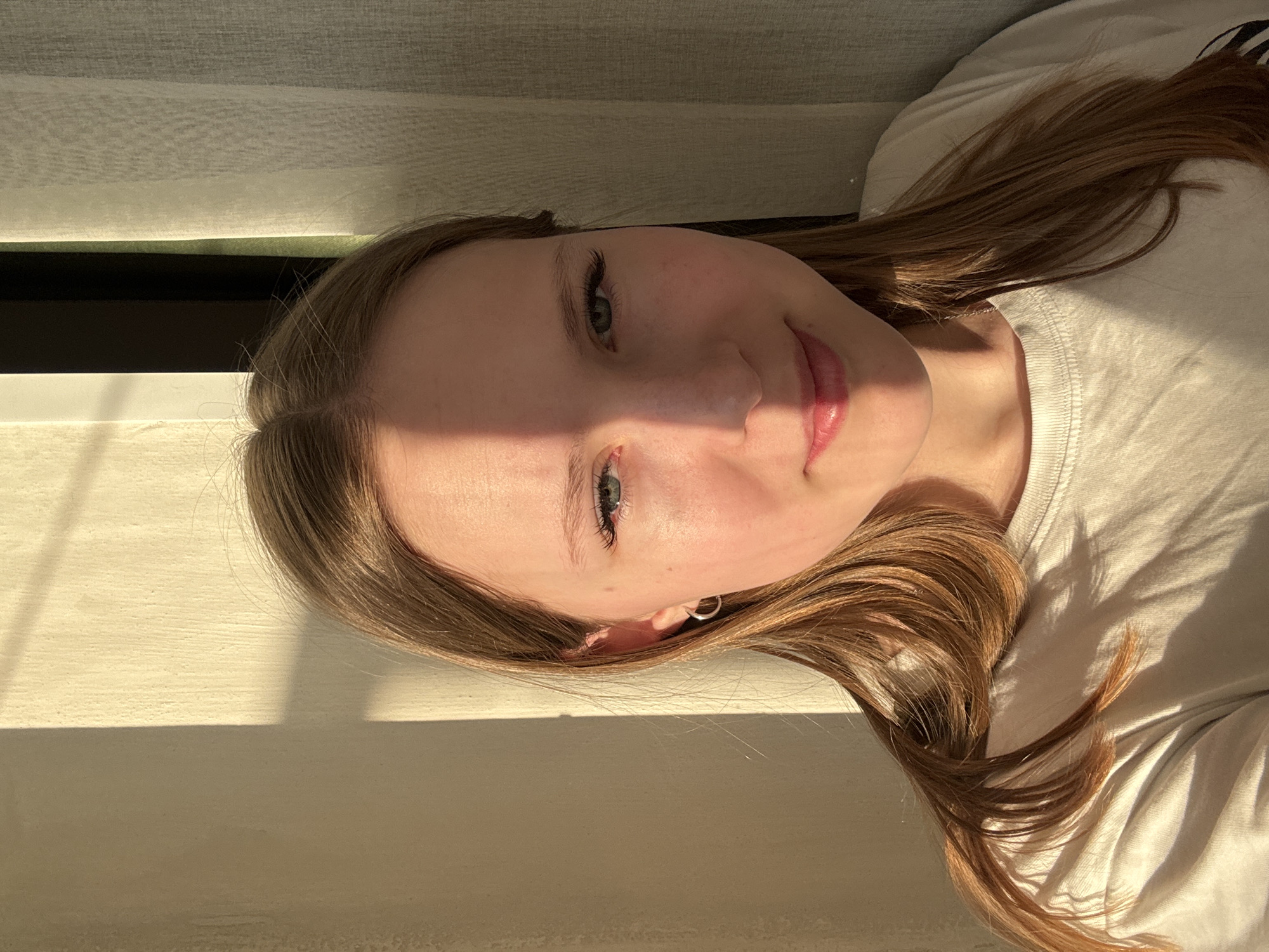 A close-up portrait shows a young individual with long, light brown hair, light skin, and blue-green eyes. The subject is positioned as if lying down or reclining, with their head tilted, looking directly at the camera with a subtle, gentle smile. They are wearing a light-colored, possibly white, crew-neck t-shirt, and a small silver hoop earring is visible in their right ear.
The scene is indoors, brightly lit by strong, warm sunlight, likely from a window. This light creates distinct, long shadows across the subject's face and the background, suggesting the time of day is late morning or early afternoon on a sunny day. The background features horizontal striped patterns, consistent with window blinds or a window frame, along with textured surfaces.
The overall impression is one of calm and relaxation. The photo was taken in an indoor setting, with the provided location context being Chaikovsky, Russia. No visible text is present in the image.