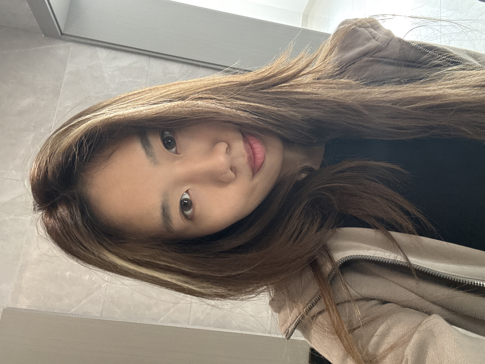 A young woman with long, light brown hair and fair skin is pictured. Her hair has a blonde highlight near her face on the left side of the frame. She is wearing a beige jacket. The background consists of light-colored wall tiling, possibly indicating a bathroom or indoor space. The location within the city of Nan'an District, China, cannot be determined from the visual information provided.
