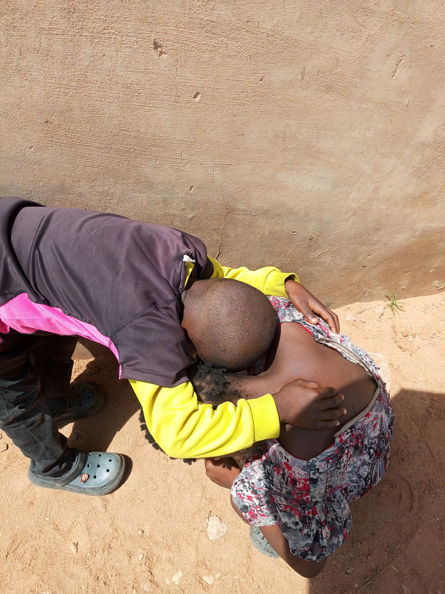 The image depicts two individuals in what appears to be an outdoor, urban environment in Tempure, Nigeria. One individual, wearing a black long-sleeved shirt with pink accents and dark pants, is bent over. Their right arm is around the shoulders of a second individual, who is kneeling and leaning forward, resting their head and upper body on the first individual's shoulder. The kneeling individual is wearing a brightly colored dress and a yellow long-sleeved shirt. The ground is a mix of sand and small rocks, and a textured, light brown wall forms the background. The scene suggests an interaction of comfort or support.