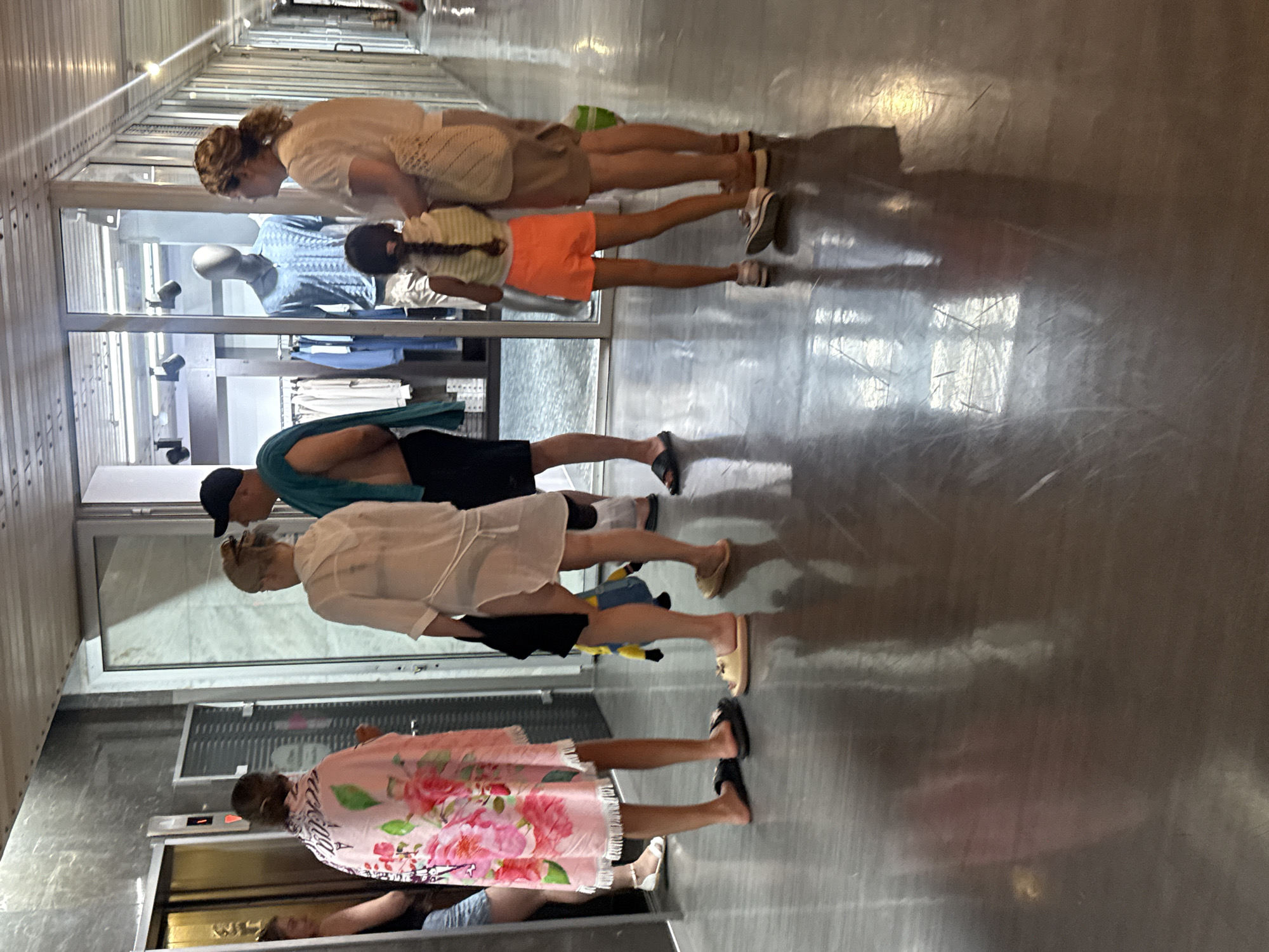A group of people are standing in a hallway with a polished floor. To the left, an elevator door is open, and a woman wearing a pink floral robe is facing it. Next to her, a woman in a white sheer cover-up and black shorts is standing with a yellow Minion toy. A man wearing a black baseball cap and a teal towel around his neck stands behind them. In the center of the frame, a young girl with braided hair is wearing an orange skirt and a striped top. She is standing in front of a store window displaying mannequins. To her right, a woman in a beige top and shorts is looking towards the store. The hallway appears to be indoors, possibly in a hotel or resort, with a store on one side and an elevator on the other. The lighting is a mix of overhead fluorescent lights and the light from the store. The floor has a reflective surface, showing distortions of the people and the environment. There is a partial view of the city of Yalta, Ukraine, indicated by the context of the location.