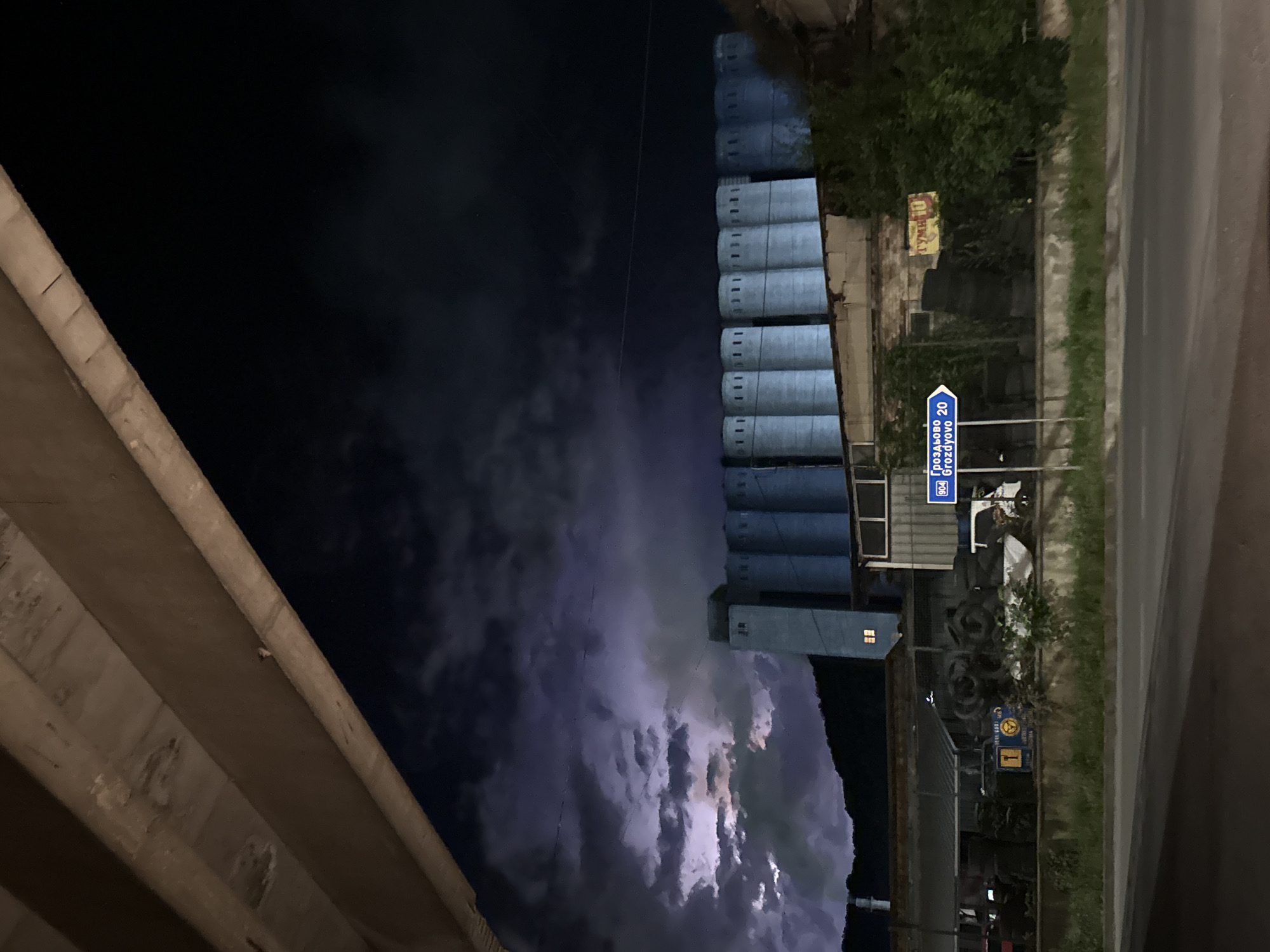 The image depicts a nighttime scene in the city of Provadia, Bulgaria, characterized by an industrial landscape under a stormy sky. In the foreground, a road is visible with a grassy median. Beyond the road, a row of large, cylindrical silos, painted light blue, dominates the middle ground. The silos have regularly spaced vertical slit windows along their upper sections. A dark, undulating mountain range forms the backdrop. The sky is dark and dramatic, with visible lightning illuminating the clouds, suggesting an ongoing thunderstorm.
In front of the silos, various industrial elements are present. These include stacks of old tires, some sheds, and a white truck. A blue directional sign is mounted on a pole, indicating "Grozdyovo 20" in both Cyrillic and Latin script. A yellow sign with red lettering, possibly advertising a business, is visible on a brick building to the right of the silos. The overall atmosphere is one of industrial activity juxtaposed with the raw power of nature. There are no people visible in the image. The lighting suggests it is night or dusk, with artificial lights from the industrial structures and natural light from the lightning.