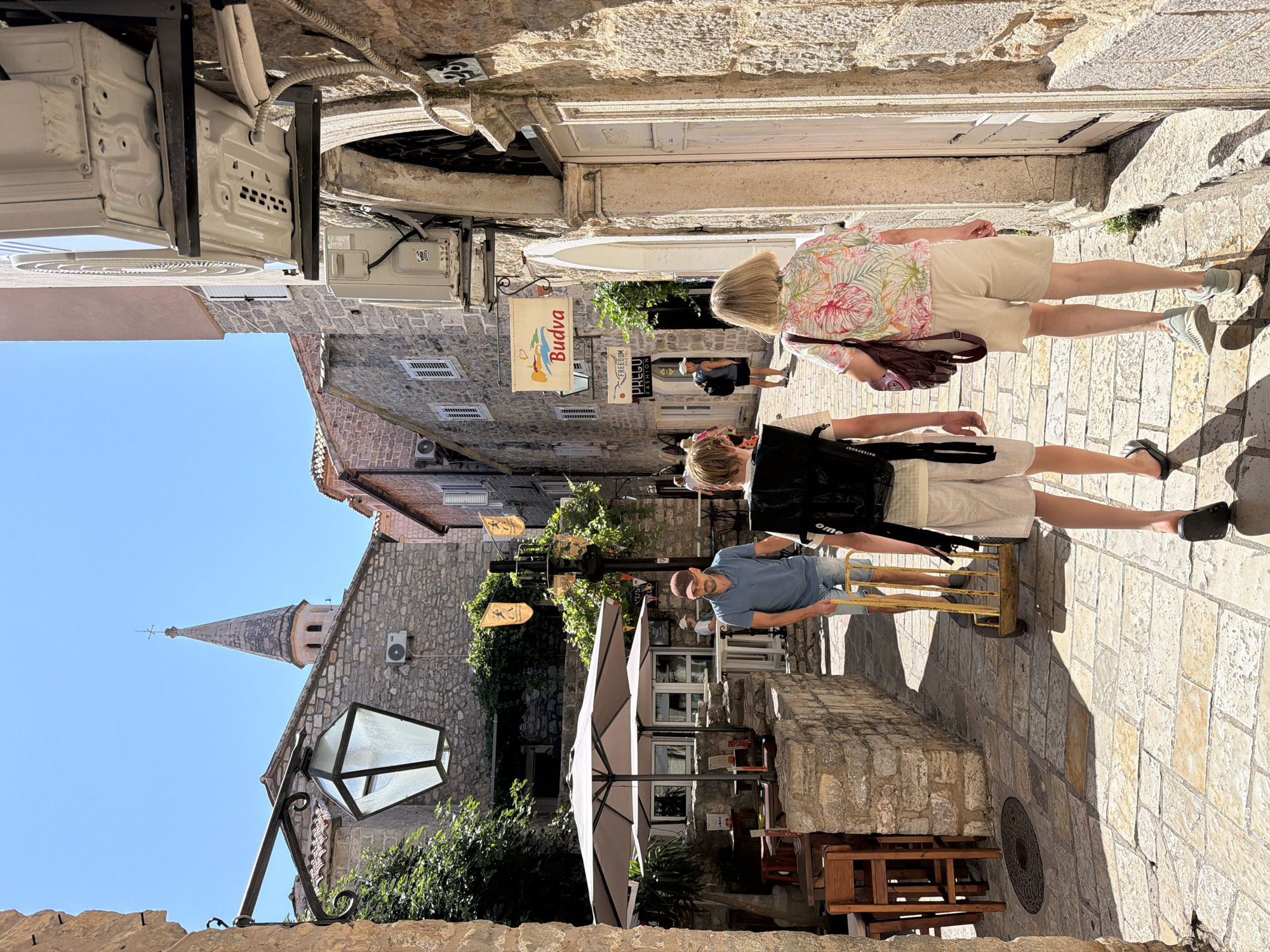The image depicts a narrow, cobblestone street in the Old Town of Budva, Montenegro. The architecture is characterized by stone buildings with terracotta roofs. The sky is clear and blue, indicating a sunny day.
In the foreground, two individuals, a younger person and an older person, are walking away from the camera. The younger person is wearing a black backpack and light-colored shorts. The older person is wearing a floral print shirt and beige shorts.
Further down the street, a man in a blue polo shirt and denim shorts is pushing a yellow wheeled cart. He is walking towards the foreground.
On the left side of the street, there is an outdoor seating area with tables and chairs, partially covered by umbrellas. A stone wall with greenery growing on it separates the street from the seating area. Above the seating area, a large, old-fashioned lantern is mounted on a wall.
On the right side of the street, there are stone buildings with wooden doors and windows. Air conditioning units are mounted on the exterior walls of some buildings. A sign with the word "Budva" is visible hanging from one of the buildings. In the distance, the spire of a church is visible above the rooftops.
The overall scene portrays a lively street scene in a historic Mediterranean town.