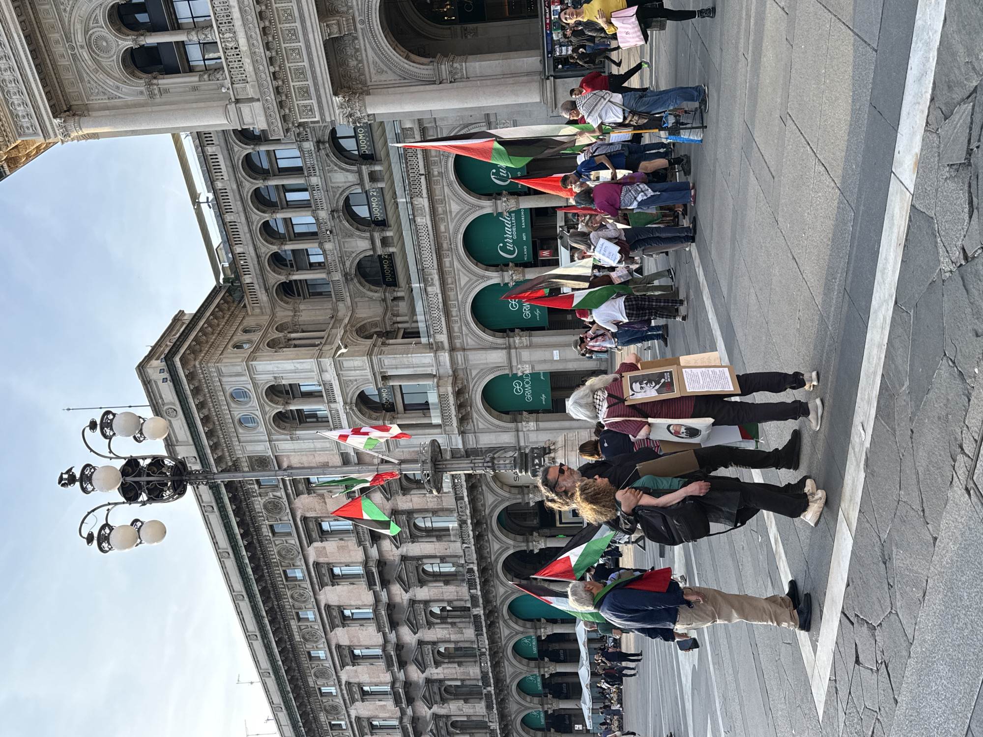 The image shows a political demonstration in Milan, Italy. A line of protesters stands on a gray stone-paved plaza, holding Palestinian flags. Some carry signs. The buildings in the background have multiple floors with rows of arched windows. Shopfronts with green signage are visible. A tall, multi-bulb lamppost stands at the edge of the crowd. The sky is visible in the upper left of the image. The buildings are ornately decorated. The text "DUOMO" and names of shops is visible on the building facades.