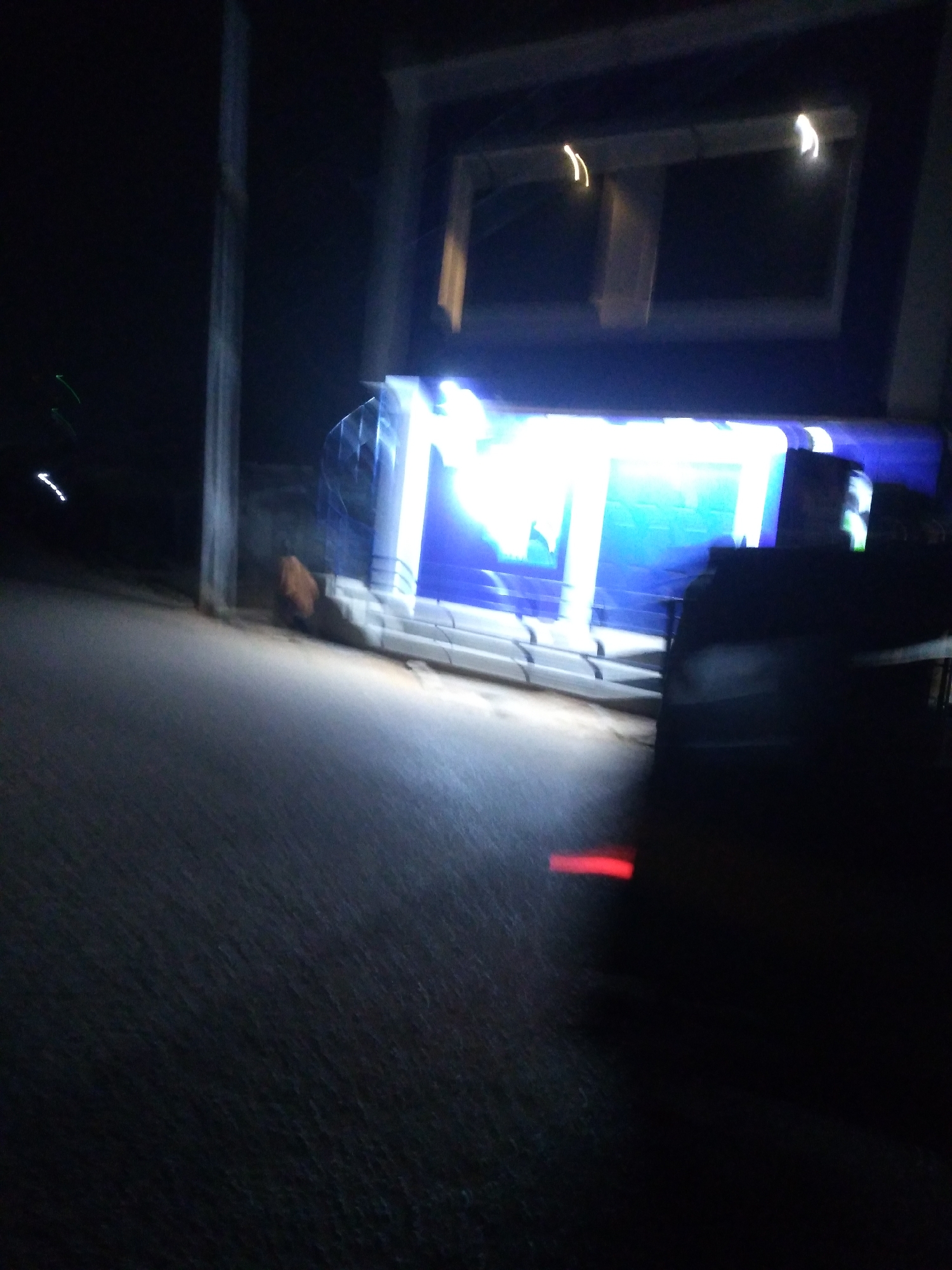 A blurry, dark image captures a scene at night. In the background, a brightly lit billboard with blue and white lettering is visible, likely advertising something. Above the billboard, a rectangular structure with yellow lights is faintly discernible. In the foreground, a dark, silhouetted object, possibly a vehicle, is parked on a paved surface, with a red light glowing on its rear. The surrounding environment is dark and indistinct, suggesting an urban setting in the city of Jos, Nigeria, likely a roadside area with commercial signage. The overall impression is one of a dimly lit, potentially commercial area at night, with limited visibility due to the darkness and motion blur.