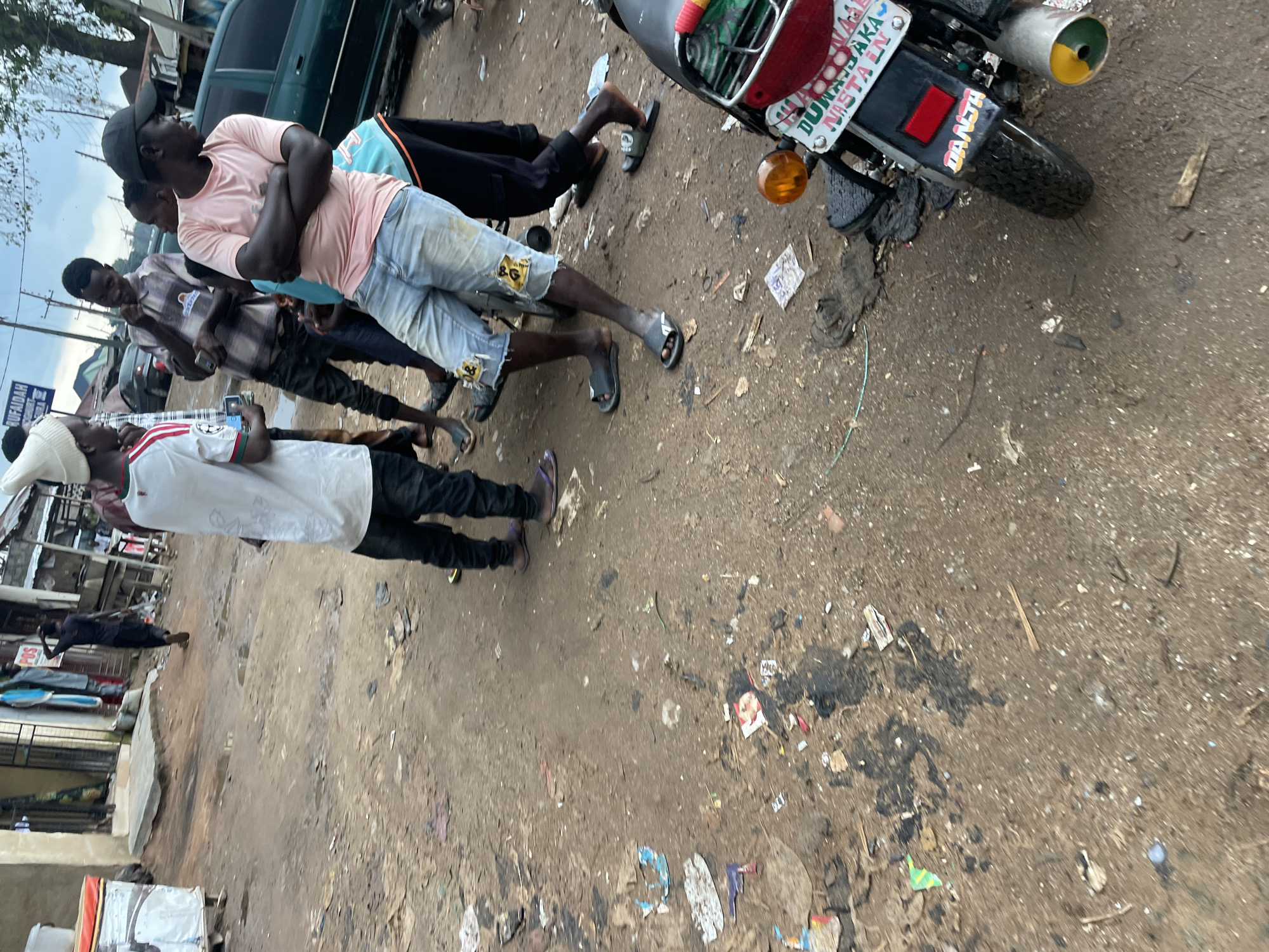 The image captures a street scene in Abuja, Nigeria, characterized by a dusty, uneven dirt road littered with various debris, including plastic wrappers, paper scraps, and what appears to be oil stains. In the foreground, the rear of a motorcycle is visible, featuring a license plate with the inscription "DUNJAKA MISTAIN" and a sticker that reads "DANJH". The motorcycle has a red taillight, a yellow-orange indicator light, and a silver exhaust pipe with green and yellow accents.
A group of young men are gathered around the motorcycle and on the road. One individual in a pink t-shirt and denim shorts is standing with his arms crossed, looking to the right. Another person, wearing a white beanie, a white t-shirt with red stripes, and dark pants, is standing in the left-center of the frame, facing left. Behind him, a man in a checkered shirt is looking down at his phone, and another person in a dark t-shirt stands to his left. Further in the background, a lone figure in a dark t-shirt and pants walks away from the camera, his back turned. A green sedan is parked on the right side of the road. The environment suggests a commercial or residential area, possibly a market or a busy street with local businesses. The overall impression is one of everyday life in an urban setting with moderate levels of untidiness.