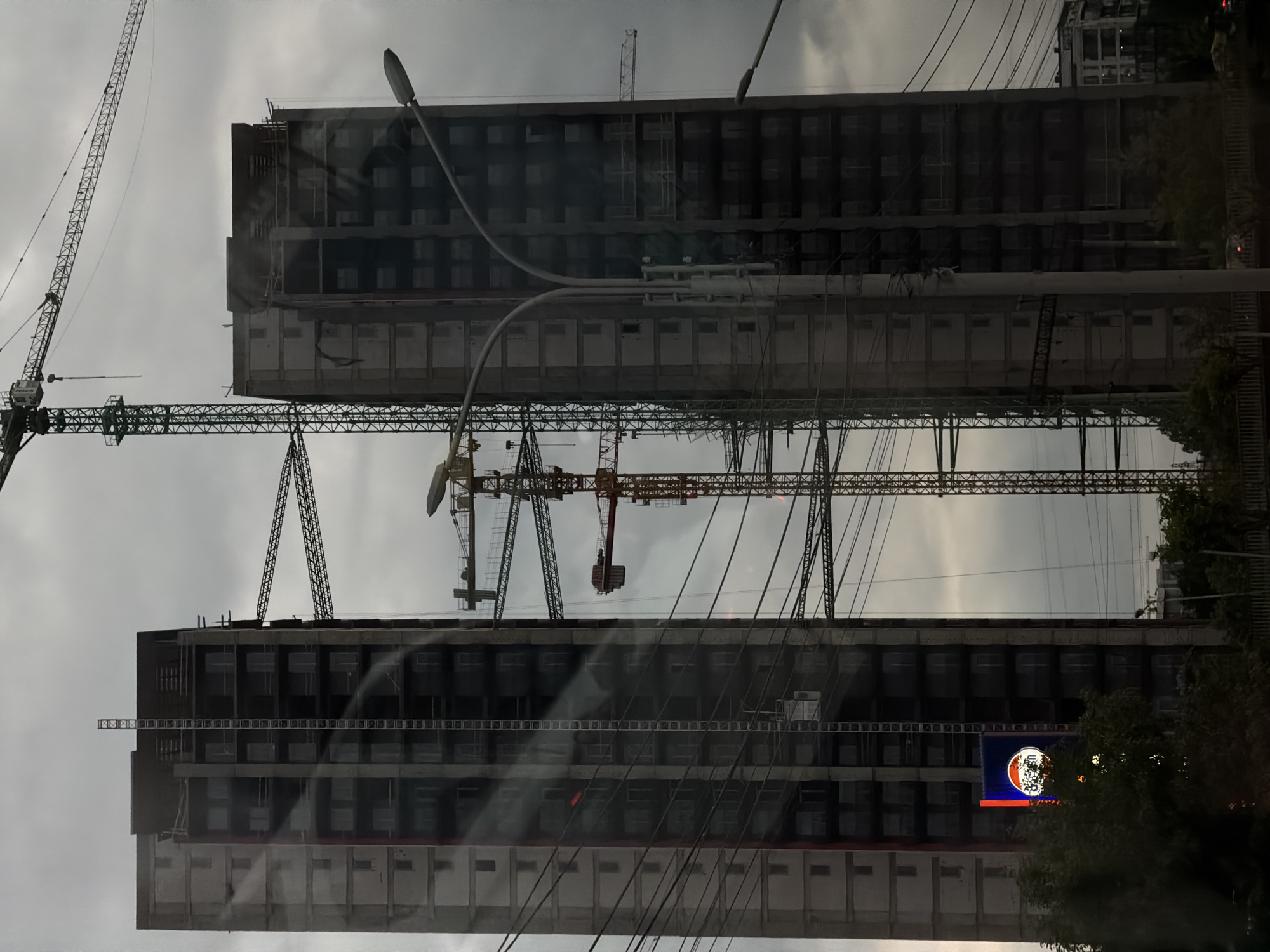 The image shows two unfinished high-rise buildings under construction in Tbilisi, Georgia. Several construction cranes are visible around the buildings. Power lines intersect the view. The sky is overcast. A tree is visible on the right. A lit sign or screen is visible on one of the buildings. The image appears to be taken from inside a vehicle, possibly a bus or car, given the reflections on the window.
