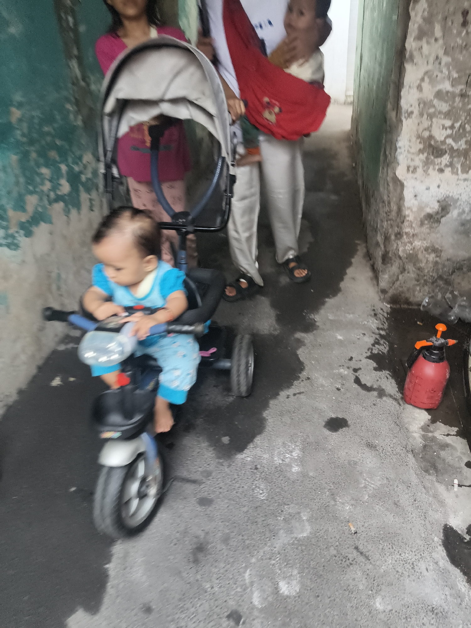 The image shows a narrow outdoor alleyway, likely in South Tangerang, Indonesia, with a young child sitting on a blue and black tricycle. The child, wearing a blue t-shirt and patterned blue pants, appears to be around one year old. Behind the child, an adult is partially visible, holding another infant in a red sling. A baby stroller is positioned next to them. The alley walls are weathered and painted in muted colors, with visible wear and tear. The ground is wet and appears to be concrete. In the background, a red spray bottle with an orange nozzle rests on the ground. A discarded cigarette butt is also visible on the wet ground. The lighting suggests an overcast day.