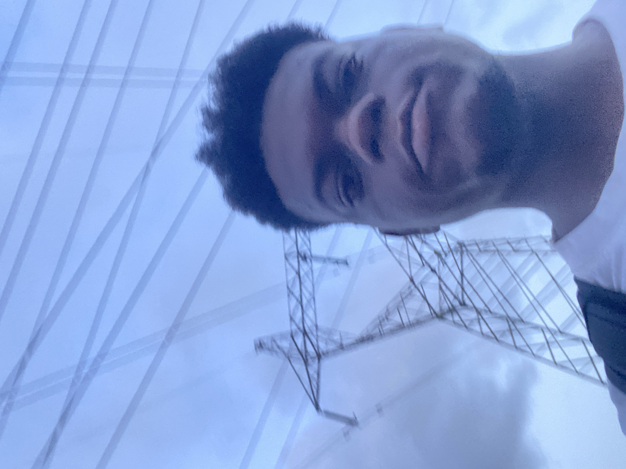 An upward-angled selfie captures a young man against a pale blue-grey sky, framed by electrical infrastructure. The man, with dark, short curly hair and some facial hair, is positioned on the right side of the frame, looking downwards with a slight, neutral expression. He is wearing a white shirt, with a dark strap visible over his right shoulder.
In the background, under a bright, diffused sky that suggests daytime, are the distinctive lattice structures of at least two large electrical transmission pylons. These metal towers are visible in the lower and mid-sections of the image. Crisscrossing across the sky, particularly in the upper and left portions of the frame, are multiple faint, linear patterns that resemble power lines or reflections, creating a layered and somewhat abstract visual effect. The overall color palette is cool, dominated by blues and greys. No text is visible.