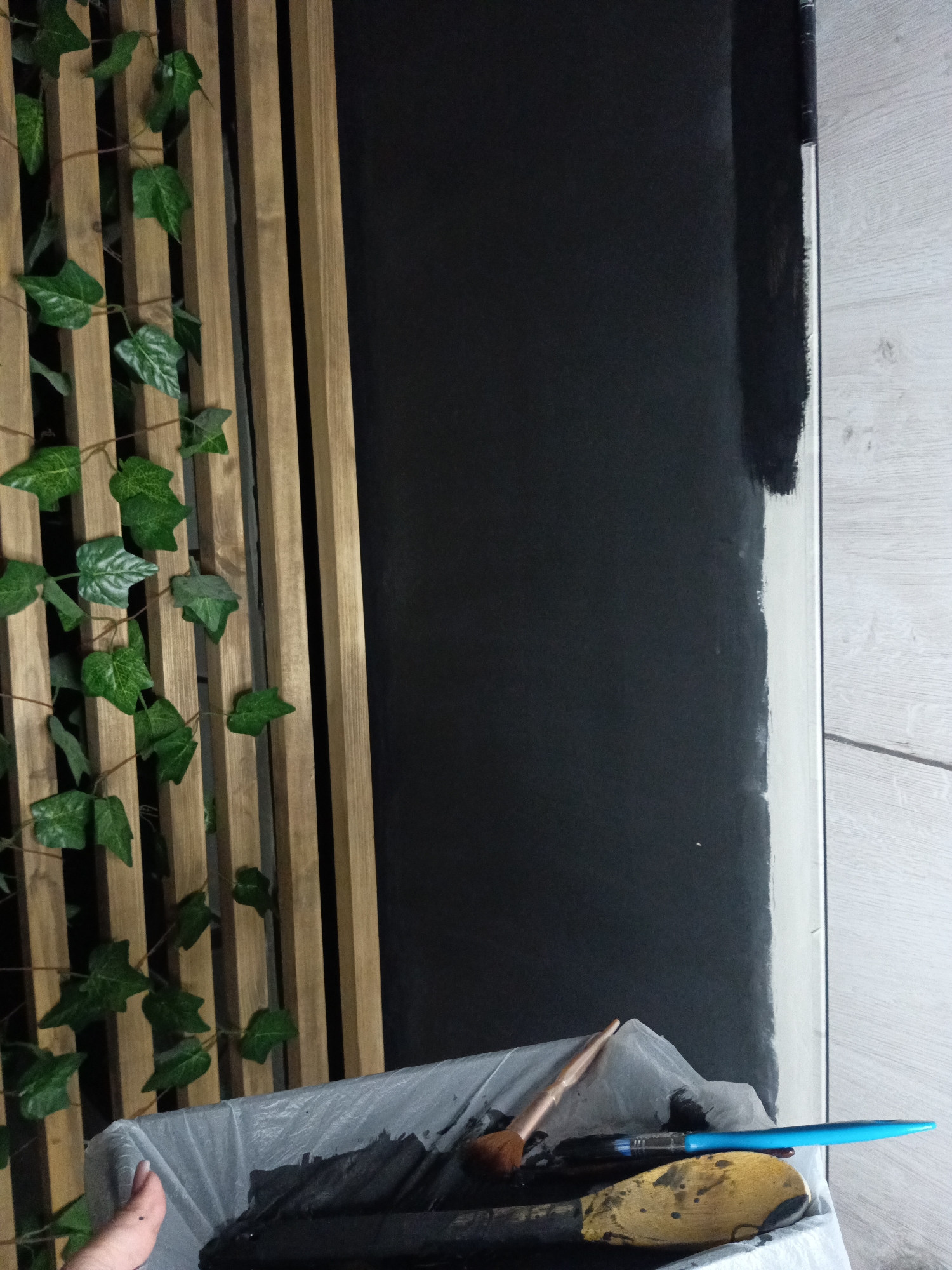 The image presents an indoor scene depicting a wall being painted black. On the left, a decorative element composed of vertical wooden slats is adorned with artificial green ivy, creating a naturalistic accent. To the right, a large section of a wall has been painted a solid black, contrasting with an unpainted or lighter wall segment on its far right edge, where the black paint shows an irregular, unfinished line, indicating an ongoing painting process.
In the lower foreground, a white plastic-lined container or bucket holds various painting tools. These include a blue-handled paintbrush, a brown-handled brush, and a wooden spoon or spatula, all stained with black paint, suggesting they are actively being used for the task. A hand, likely belonging to the person undertaking the painting, is partially visible on the bottom left, reaching into the container. The floor visible on the right is composed of light-colored wooden planks or laminate. The overall setting suggests a home renovation or decoration project taking place in Фоки, Russia. There is no visible text.