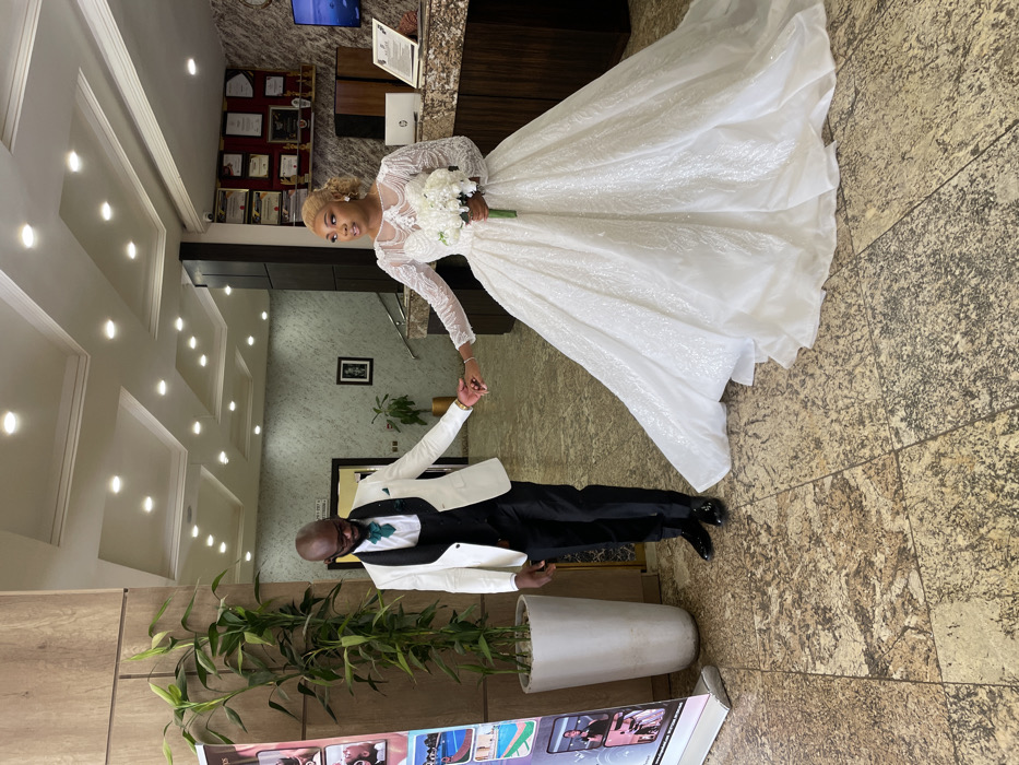 The scene depicts an indoor public space, likely a hotel lobby or event venue, located in Barakin Lerawi, Nigeria. Two individuals, a male and a female, stand centrally, holding hands. The male, with dark skin and a bald head, wears glasses, a white tuxedo jacket with black lapels, a dark green vest and cravat over a white shirt, black trousers, and shiny black dress shoes. He looks downward and to his right. The female, with lighter skin and blonde curly hair, wears an embellished white long-sleeved ball gown wedding dress and holds a white floral bouquet in her right hand. She looks forward with her mouth slightly open. The floor is tiled with a mottled brown and beige pattern. To the left, a tall green plant in a white cylindrical planter stands next to a colorful vertical banner. In the background on the right, a dark wooden reception desk with a light-colored countertop features a laptop and framed certificates on the wall above it. A wall-mounted screen displays an underwater scene. The ceiling is white with multiple recessed circular lights, illuminating the area.