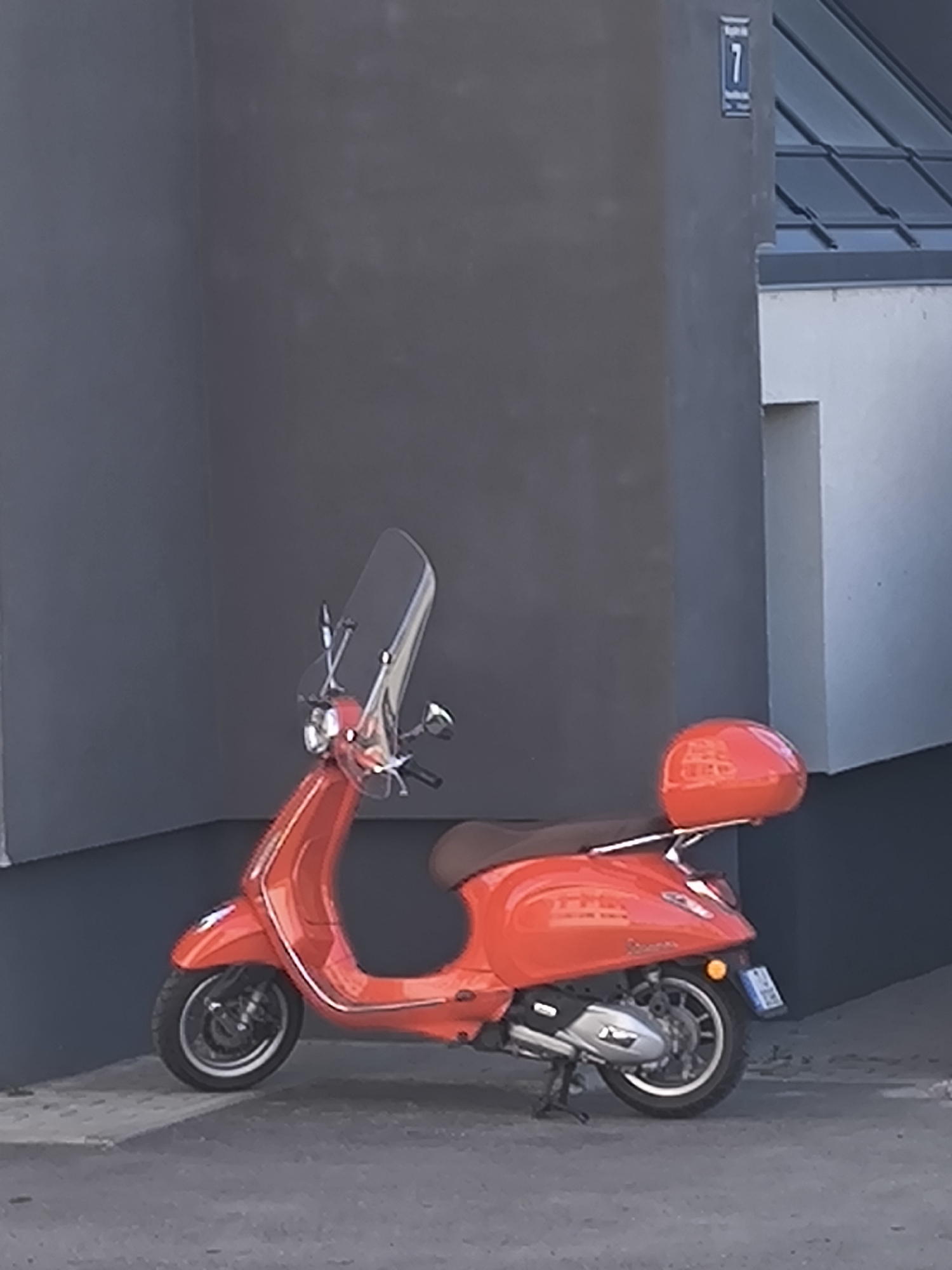 An orange Vespa scooter is parked on a paved surface next to a grey building. The building has a number "7" on its corner. The architecture suggests the location is in Riga, Latvia. The scooter has a windshield and a rear storage container.