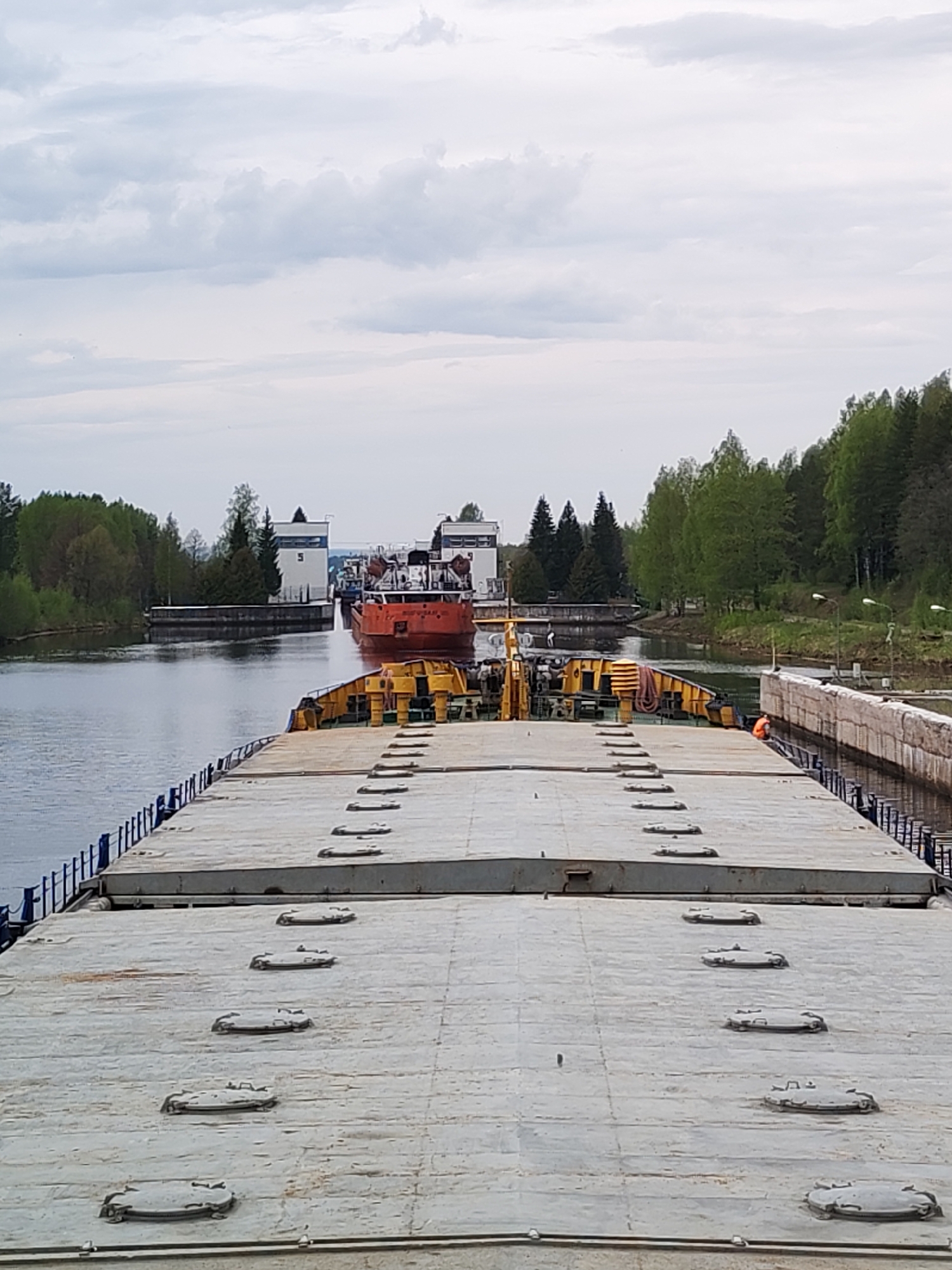 The image depicts a barge navigating a canal near Alekseevskoe, Russia. The foreground features the deck of a barge with numerous hatch covers arranged in parallel lines. In the midground, another orange vessel is seen moving through a canal lock with building '3' on one side and building '5' on the other. The surrounding environment consists of green trees and vegetation lining the canal banks, and an overcast sky suggests daytime. The lock mechanism and concrete retaining walls are visible on both sides of the canal. A person wearing a bright orange shirt is near the wall.