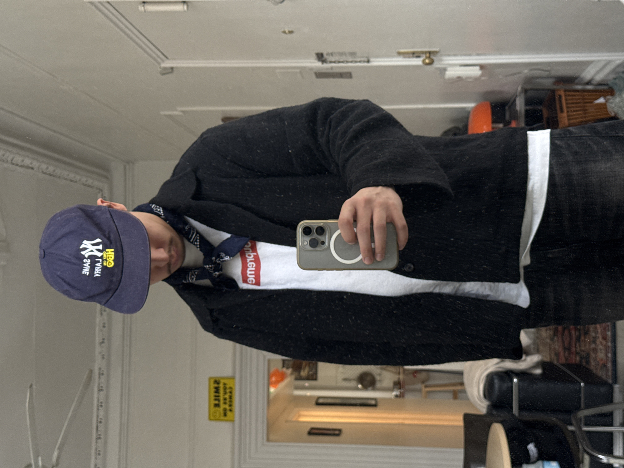 A male subject is depicted in a mirror selfie taken within an interior residence, located in the city of New York, United States of America. The subject is seen from the chest up, wearing a navy blue baseball cap with a yellow New York Yankees "NY" logo, superimposed with yellow reversed text "SANE LARRY HBO". He also wears a navy blue bandana tied around his neck, a white t-shirt featuring a red box logo (reversed "Supreme"), a dark, textured, collared jacket, and dark denim trousers. He holds a light-colored smartphone, visible with a MagSafe ring on its rear, in his right hand. The reflection reveals light-colored walls with decorative crown molding and a partial view of a white ceiling fan. A patterned rug covers the floor. Visible furniture includes a portion of a round table with a dark base and a dark, possibly leather, armchair draped with a light-colored blanket. Through an open doorway in the background, framed art, a hanging pan, an orange spherical object, and a woven basket are discernible. A yellow sign with the reversed text "SMILE YOU'RE ON CAMERA" is affixed to the wall near the doorway. A white door with a brass doorknob and a white wall plate is visible on the right side of the frame.