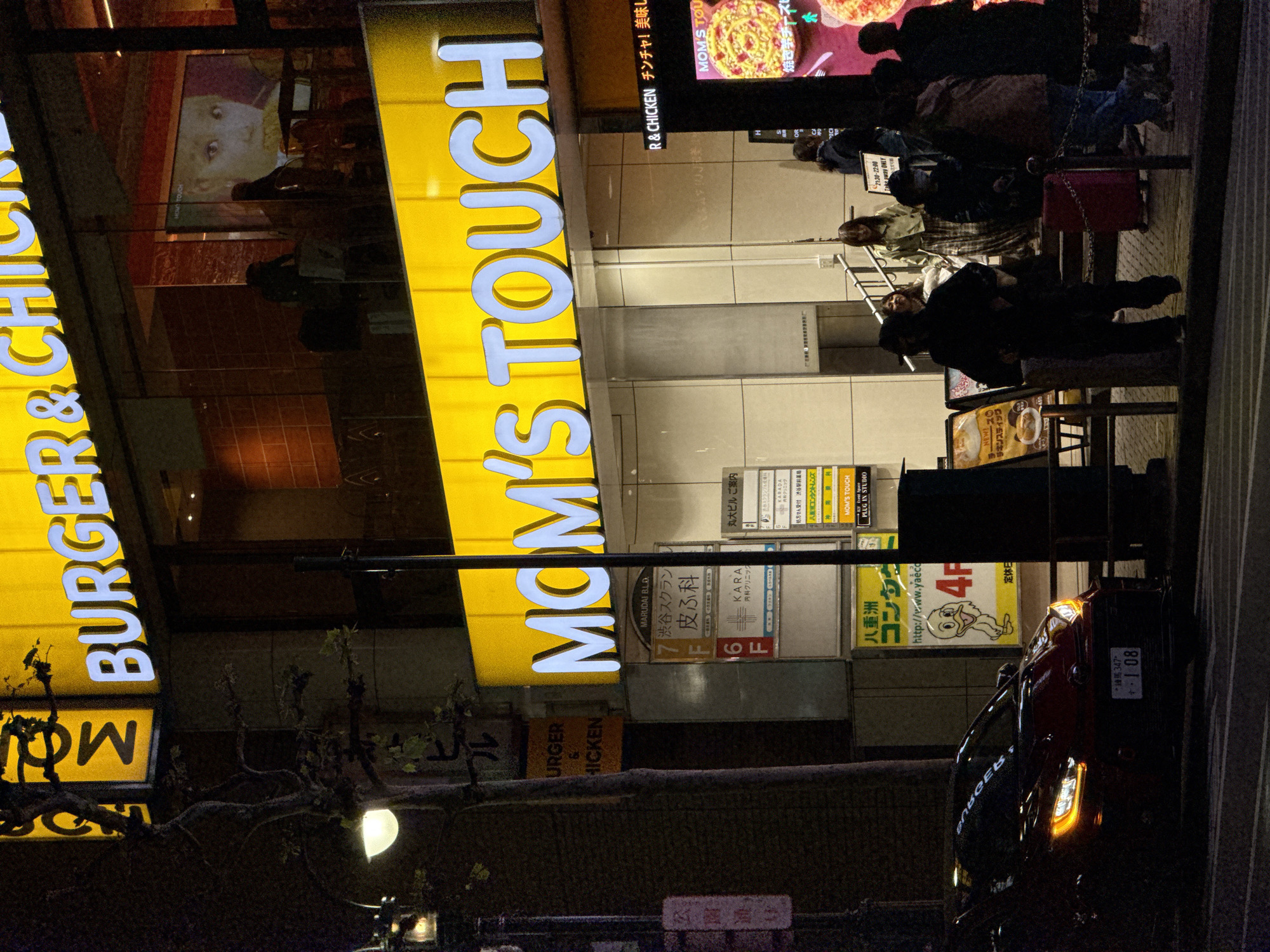 An urban street scene unfolds at night, likely in Shibuya, Japan, characterized by a brightly illuminated building housing various businesses. The central focus is a multi-story commercial building with a prominent yellow sign displaying "MOM'S TOUCH" and "BURGER & CHICKEN" in large, glowing letters. Reflections of people and interior lighting are visible in the building's glass facade.
Several people are gathered on the sidewalk in front of the building, some appearing to be queuing or waiting. Their forms are largely silhouetted against the bright signs and interior lights, making individual features indistinct.
Below the main fast-food signs, smaller signs in Japanese advertise other tenants of the "MARUDAI BLD." These include "渋谷スクラン 皮ふ科" (Shibuya Scramble Dermatology) on the 7th floor, "KARA 内科クリニック" (Kara Internal Medicine Clinic) on the 6th floor, a pharmacy ("処方せん受付 渋谷駅前薬局"), and "八重洲コンタクト" (Yaesu Contact) with a cartoon duck and web address "http://www.yaeco" on the 4th floor. Other text mentions "R & CHICKEN チンチャ! 美味し" (Really! Delicious!) and "MOM'S TOUCH PLUG IN STUDIO" alongside images of food like pizza. A "NEW! ・チキンスティック 450" (Chicken Sticks 450) offer is visible, as well as a sign indicating "21:30-22:00 Take away ONLY."
A dark-colored car, possibly red, is parked or driving on the street in the foreground to the right, with its headlights and taillights on. Its Japanese license plate is partially visible, reading "*練馬347* ・1 08". A bare-branched tree with some foliage and a streetlight illuminate the lower left corner. The overall ambiance is busy and energetic, typical of a city's nightlife.