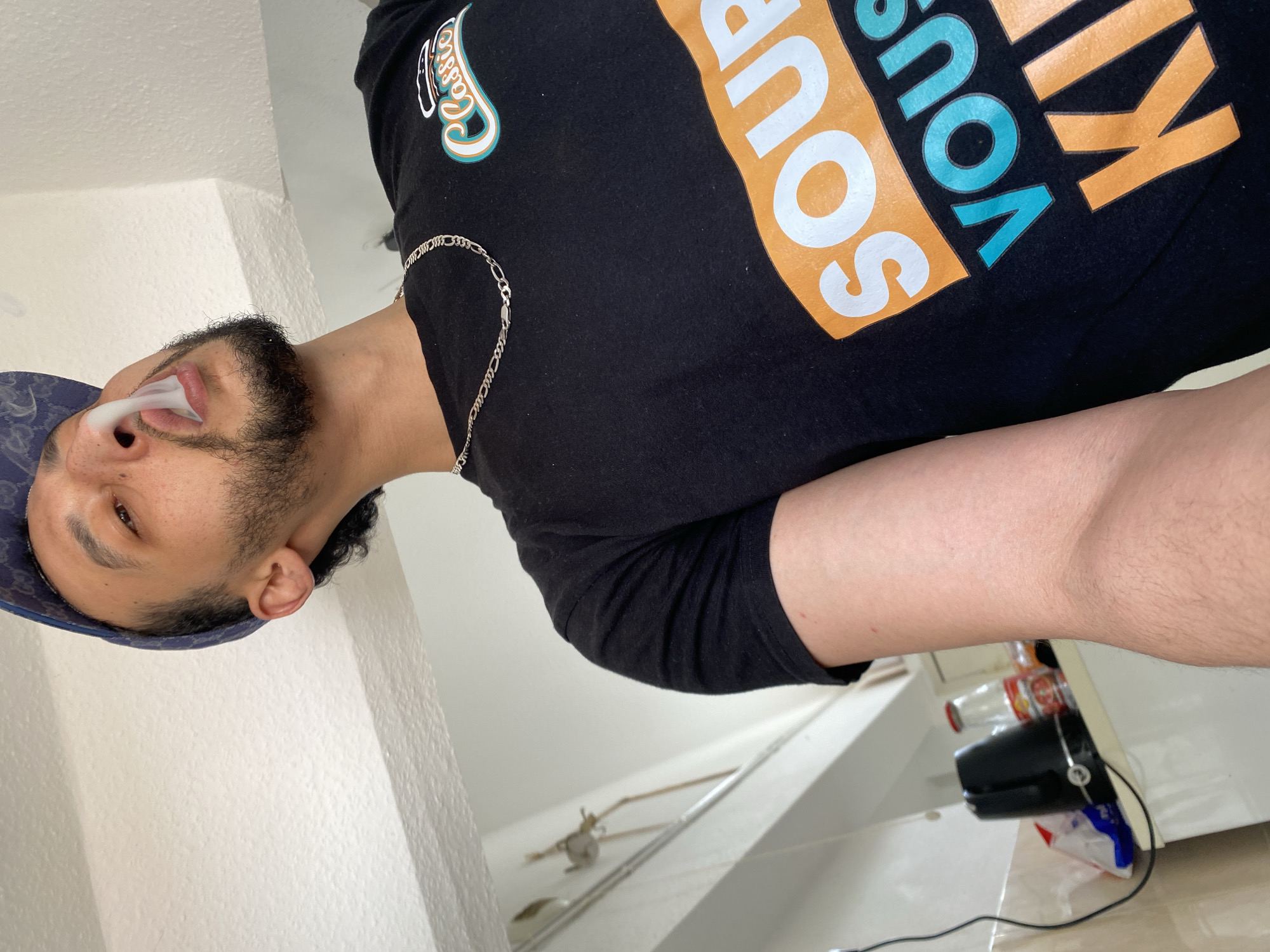 A man with a beard and a patterned baseball cap is shown in a photograph. He is wearing a black t-shirt with a graphic design that includes orange, turquoise, and white lettering. A silver chain is visible around his neck, and he appears to be exhaling smoke. The background includes a white wall, part of a sink, and various objects on a surface behind him, including bottles and what appears to be a black electronic device. The photograph has been rotated. There is no indication in the photo that this was taken in Bischheim, France.