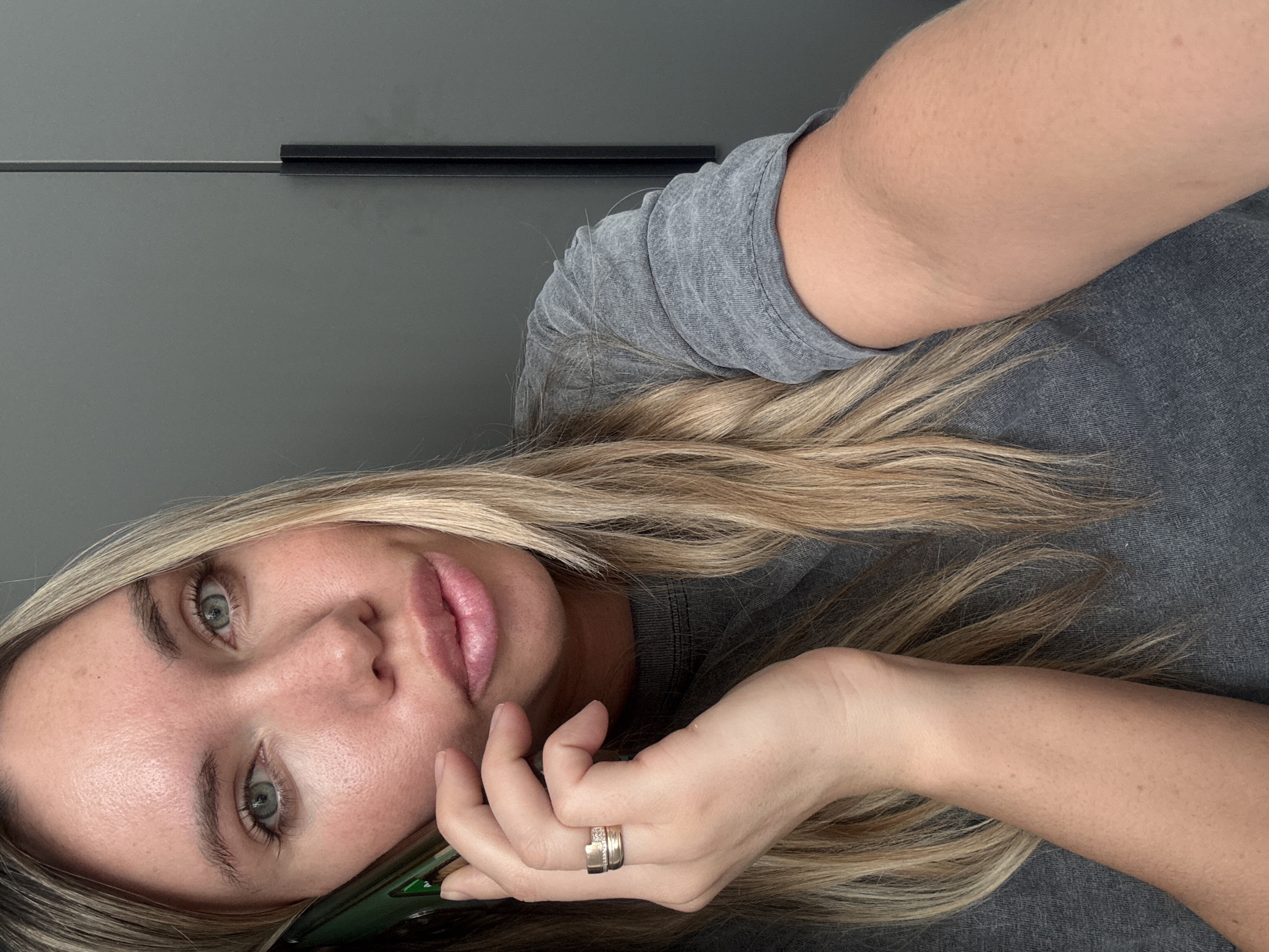 The image depicts a female individual captured from the chest up, situated indoors in a location identified as Votkinsk, Russia. She possesses long, straight blonde hair and light blue-green eyes. Her skin exhibits a fair complexion with visible pores and minor redness across the forehead and cheeks. She is dressed in a short-sleeved, grey t-shirt with a textured finish. Her left hand, adorned with a two-band gold-colored ring on the ring finger, holds a dark green smartphone to her left ear. Her gaze is directed forward, towards the camera, and her lips are slightly parted. The background consists of a plain, dark grey surface, likely a wall or cabinet, featuring a vertical dark trim or handle on the right side of the frame. Specific interior details within Votkinsk are not visually ascertainable from the image.
