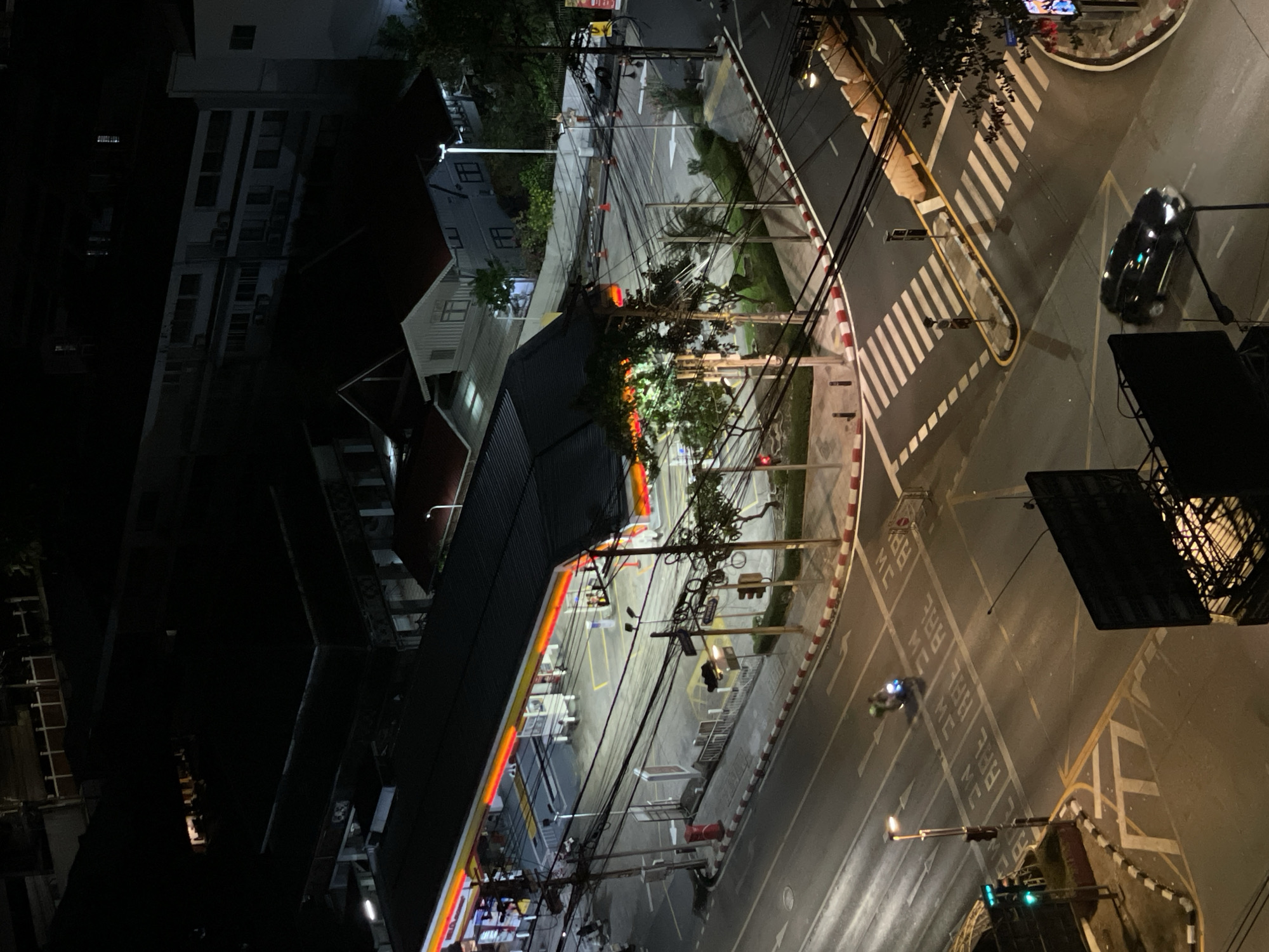The image depicts a nighttime scene in Bangkok, Thailand, taken from an elevated perspective. The primary focal point is a brightly illuminated Shell gas station with an orange and yellow canopy situated at a T-intersection. The street is marked with pedestrian crosswalks and motorcycle lane indicators. A few vehicles are present, including a dark-colored car and a scooter. Several buildings of various heights surround the intersection. Traffic lights and utility poles, along with an extensive network of electrical wires, further define the urban landscape.