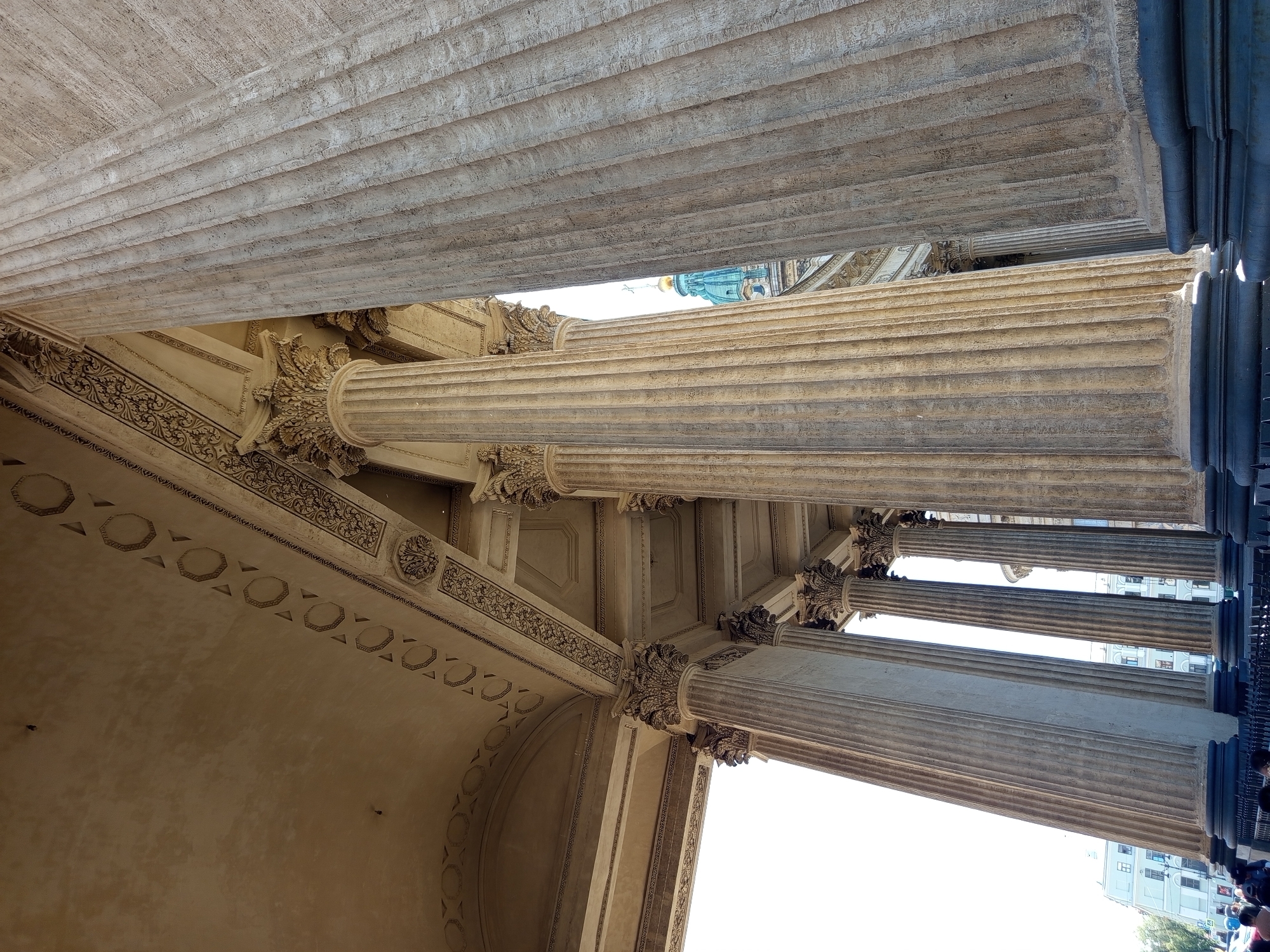 This image shows a low-angle view of the colonnade of Kazan Cathedral in Saint Petersburg, Russia. The massive stone columns are fluted and topped with ornate capitals. The ceiling above the colonnade features decorative friezes with geometric patterns and floral motifs. In the background, a portion of the cathedral's green dome with a cross is visible. The scene suggests a sunny day, with bright light illuminating the architecture. People are visible at the bottom of the image, suggesting the area is accessible to the public. The overall impression is one of grandeur and classical architecture.