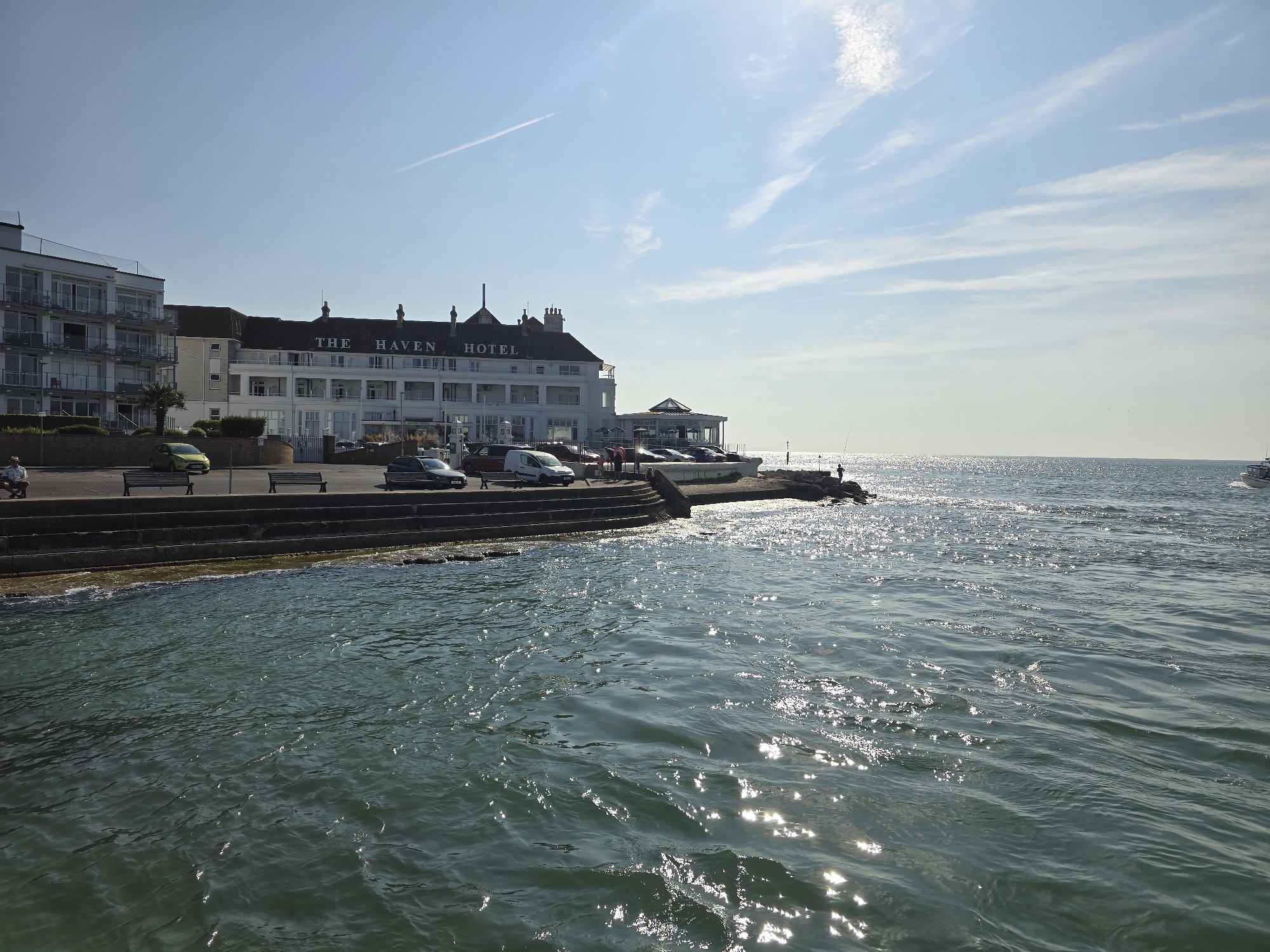 The image depicts a view of The Haven Hotel in Poole, United Kingdom, situated along a waterfront. The hotel, a large white building with multiple stories and balconies, is prominently featured. In front of the hotel, there is a promenade with benches and parked cars. The water in the foreground is a deep teal color with sunlight glinting off the surface, indicating a sunny day. There are some steps leading down to the water. On the far right, a small boat can be seen in the distance. The sky is blue with some wispy white clouds. A person is visible standing on a rocky outcrop near the water on the right side of the image.
