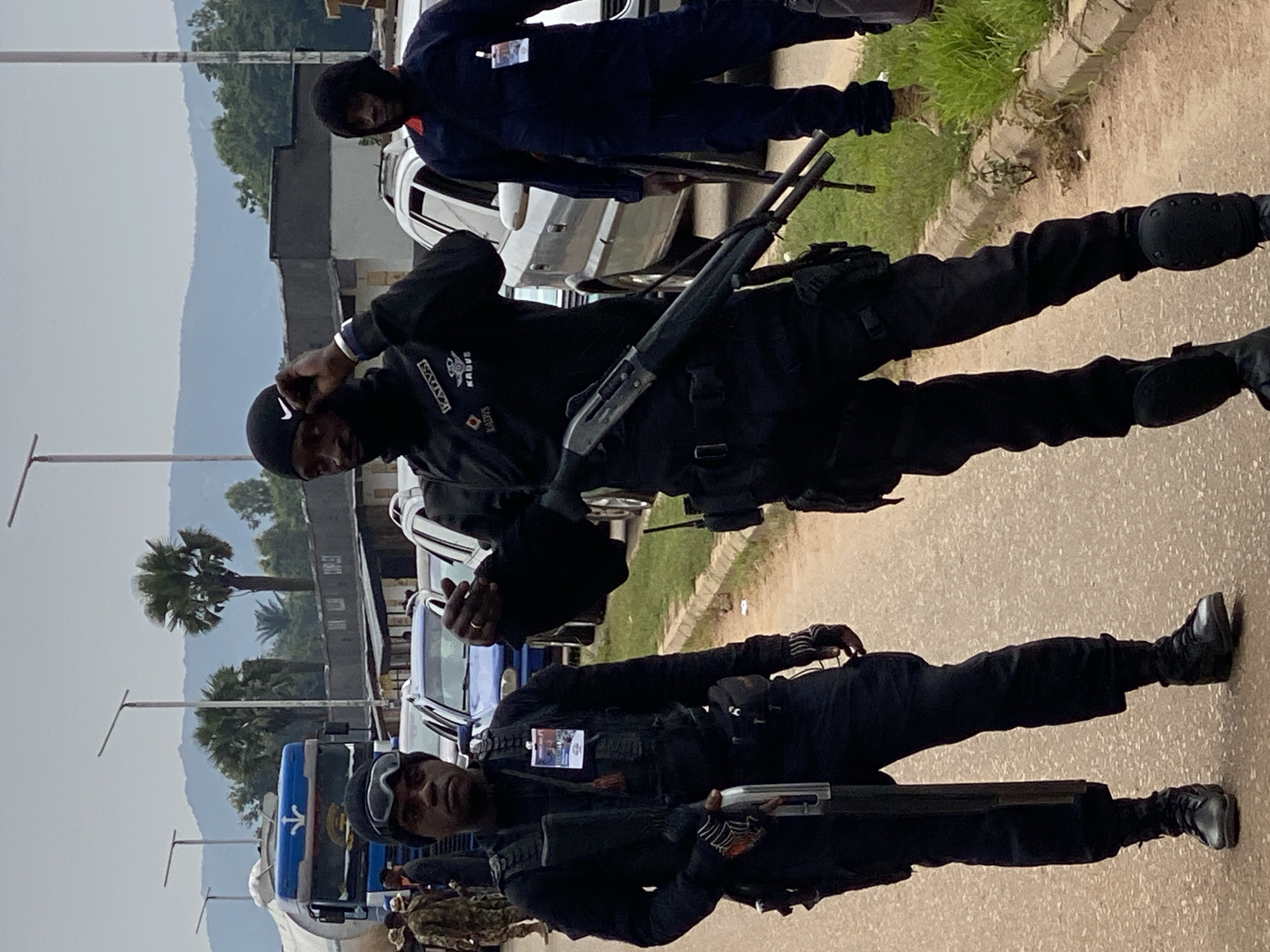 Three men stand on an asphalt road. One wears dark blue clothing. Two wear black clothing, tactical gear, and carry shotguns. Vehicles are parked in the background. Trees and distant hills are visible. This is likely in Maigizo, Nigeria.