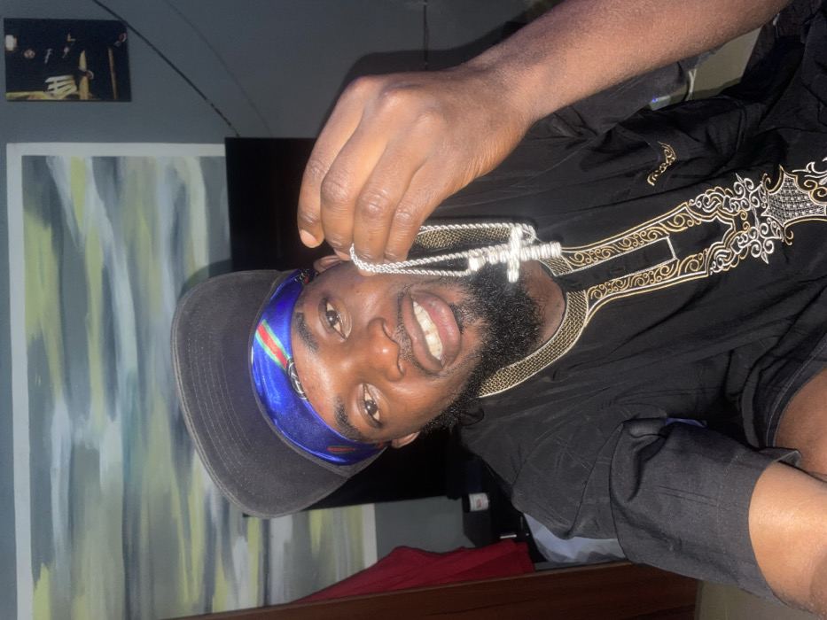 A man poses with a silver cross necklace. He has dark skin, a beard, and wears a black baseball cap over a blue and green headscarf. He is wearing a black shirt with gold embroidery. He is indoors with a painting on the wall and a small framed photograph above it. A bed with red sheets is visible below him. The image is rotated counterclockwise.