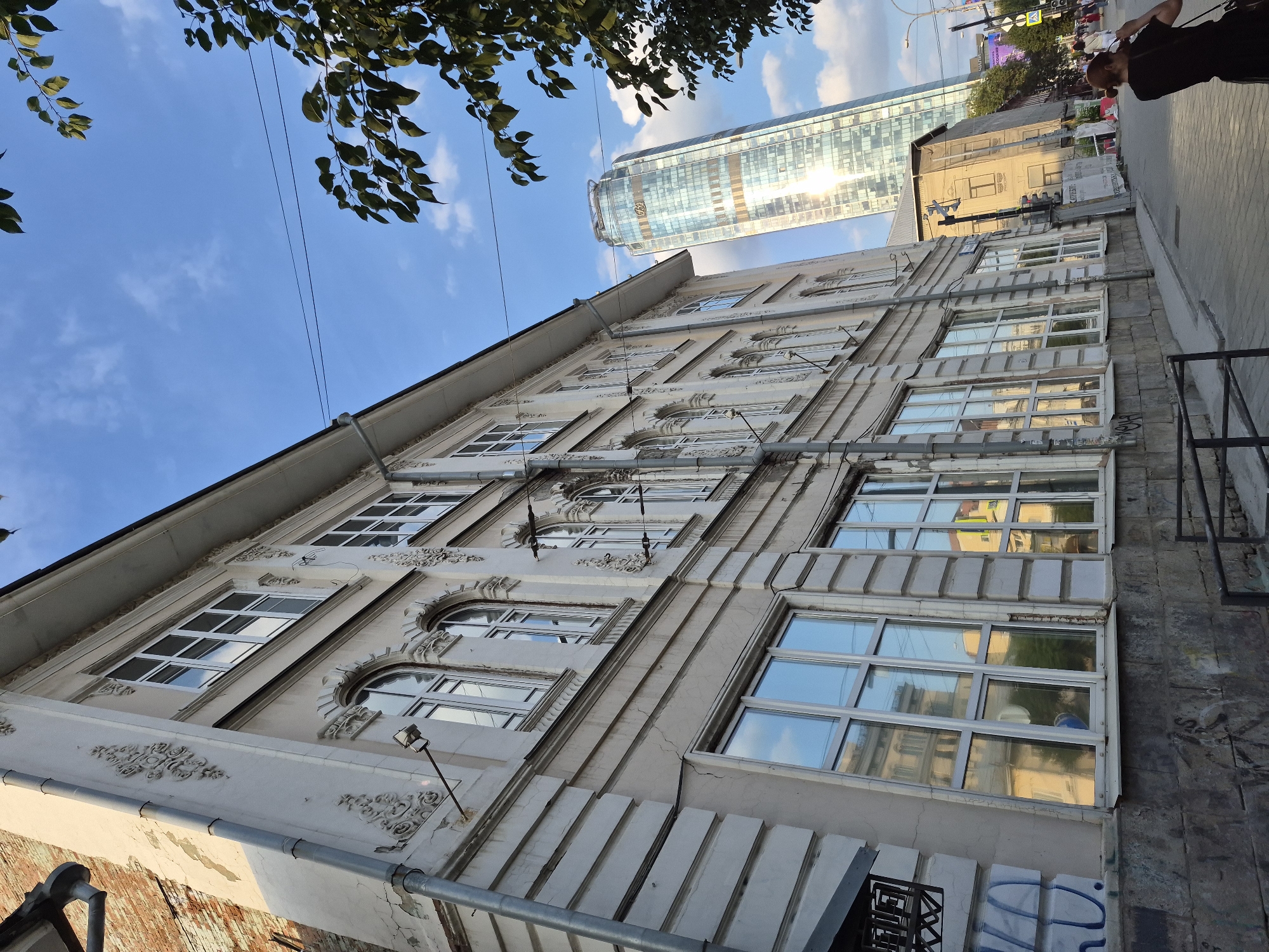 In Yekaterinburg, Russia, an old, ornate building with decorative plasterwork and large windows stands on a street. The building's facade is light-colored, with some signs of wear and peeling paint on an adjacent brick section. A modern, glass skyscraper with a distinctive curved top rises in the background, reflecting the sky and sunlight. A person is visible in the foreground, partially obscured by the frame, holding a phone to their ear. Trees with green leaves frame the scene from the right, and power lines are strung across the blue sky with scattered clouds. The street appears to be paved, and there are other buildings and street elements further down, including traffic lights and signage.