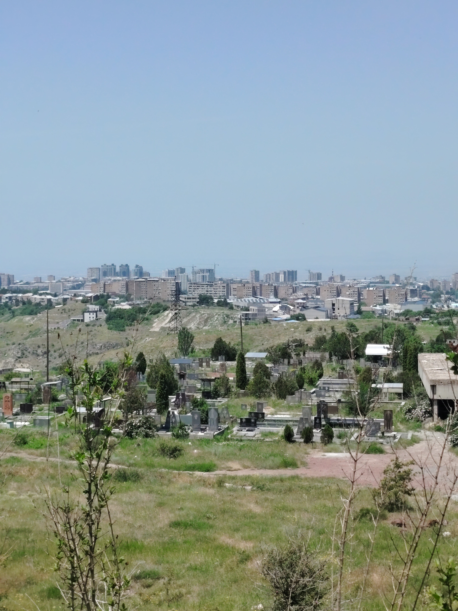 The image shows a daytime view of a cemetery in Jrvej, Armenia, with a city skyline in the background. The cemetery is filled with gravestones and mausoleums, some of which appear to be made of stone and metal. The ground is covered in grass and dirt paths. In the distance, there is a sprawling urban landscape with numerous multi-story buildings, including residential apartments and what appears to be construction sites. A large electrical pylon stands on a hill between the cemetery and the city. The sky is clear and blue. The overall scene suggests a juxtaposition of a place of remembrance with the ongoing life of a modern city.