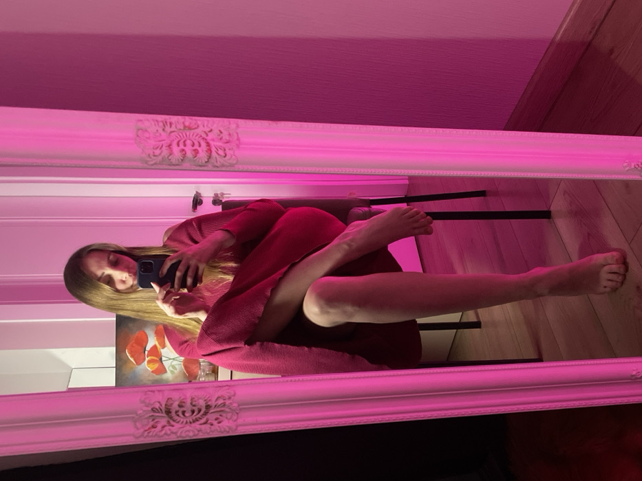 The scene depicts a female individual, likely located in a room within Batumi, Georgia, captured via a mirror selfie. The subject, with long, light brown hair, is seated on a chair, facing a large, ornate white-framed mirror. She is wearing a long-sleeved, ribbed dress or tunic in a fuchsia or deep pink color. Her left leg is crossed over her right, and both bare feet are visible. She holds a dark-colored smartphone with both hands, appearing to operate it. The room is heavily illuminated with a strong pink light, casting a uniform fuchsia hue over the visible environment. Reflected in the mirror, behind the subject, is a white door with a silver handle and an inserted key, positioned on the right side of the frame. Above the door handle, a painting depicting red poppy flowers is visible. Below the painting, a light-colored furniture piece with small objects on top is partially discernible. The floor consists of light-colored wooden planks. The wall to the right of the mirror (outside the reflection) shows a textured surface, also illuminated in pink.