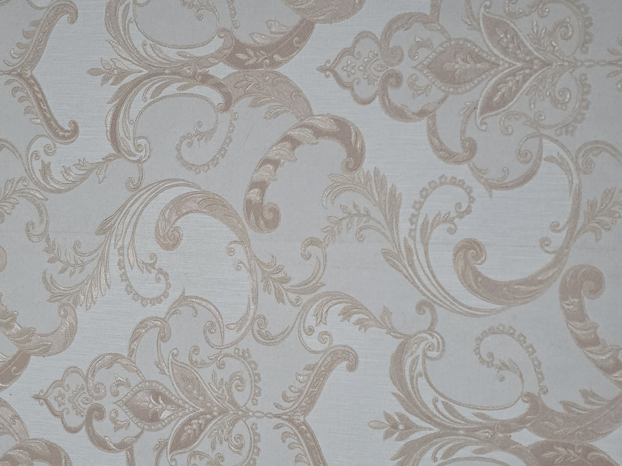 The image displays a close-up of textured wallpaper with an ornate damask pattern in shades of beige and cream. The pattern consists of elaborate floral and scrolling motifs repeated across the surface. The texture of the wallpaper is visible, suggesting a woven or embossed material. There are no people, objects, or environmental details indicating a specific location in the city of Qarabalıq, Kazakhstan. The image is purely a close-up of a decorative wallpaper pattern.