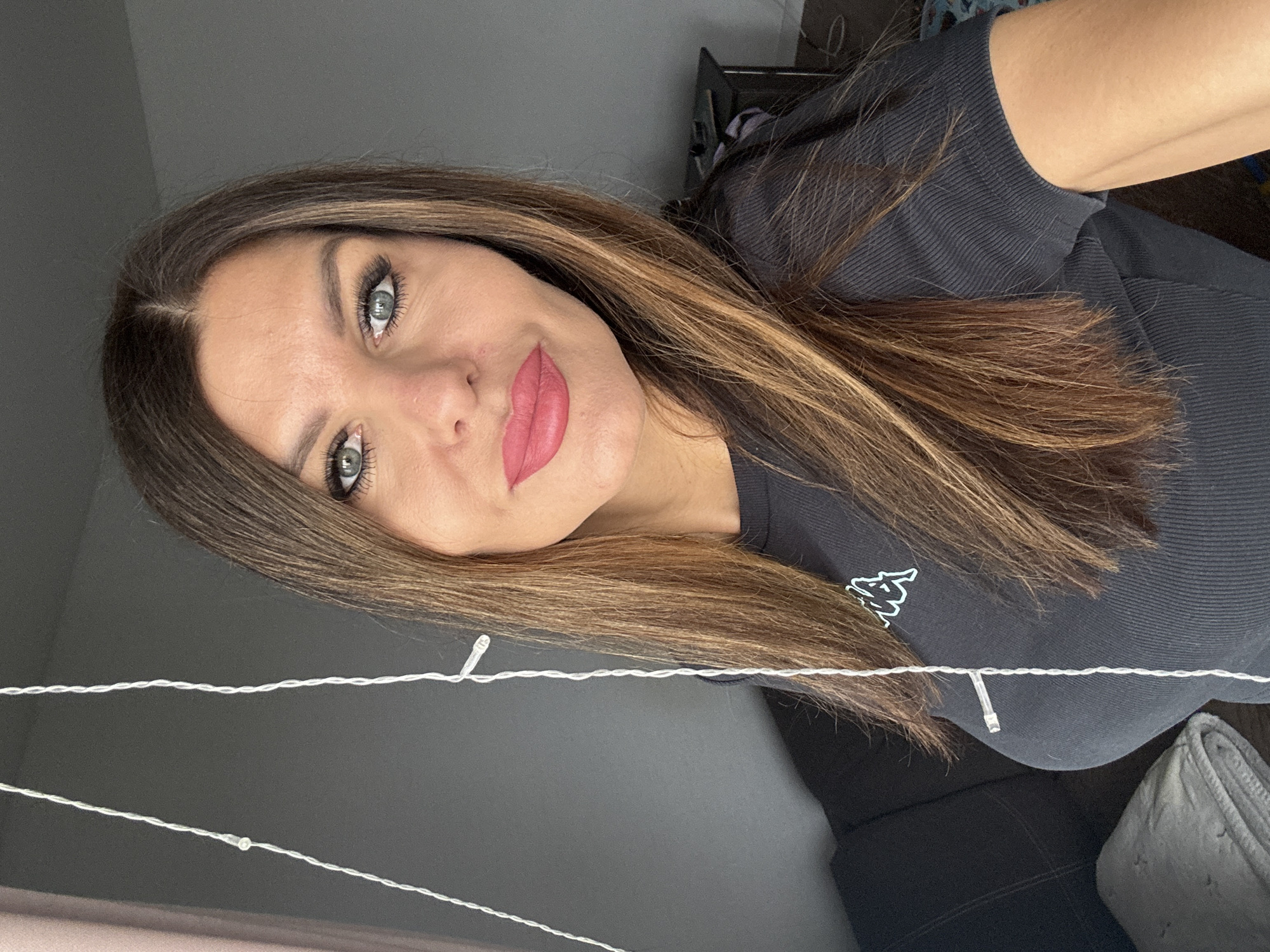 A woman is shown in a selfie-style image. She has long, brown and blonde highlighted hair and light-colored eyes. She's wearing a dark-colored, ribbed shirt with a logo on the chest. There is a string of small, white lights in the foreground, and behind her is a gray wall. A couch and a folded blanket are visible in the background. There is no information in the image to indicate this was taken in Chaikovsky, Russia.