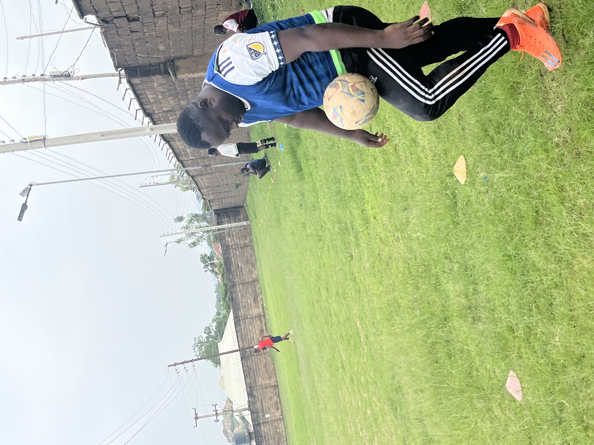 The image depicts a group of young men playing soccer on a grassy field. The primary subject is a young man in the foreground, wearing a blue and white jersey, black athletic pants with white stripes, and bright orange soccer cleats. He is actively engaged with a soccer ball, appearing to be in the process of kicking or controlling it. Another young man in a red shirt and black shorts is visible in the background, also on the field. The setting is an outdoor soccer field, characterized by well-maintained green grass. A brick wall and some electrical poles are present in the background, suggesting an urban or semi-urban environment. The sky appears overcast, indicating a cloudy or potentially rainy day. The overall atmosphere suggests a casual soccer practice or game. There is a logo on the jersey of the main subject, but the text is not legible. The image captures a moment of athletic activity and youthful energy.
