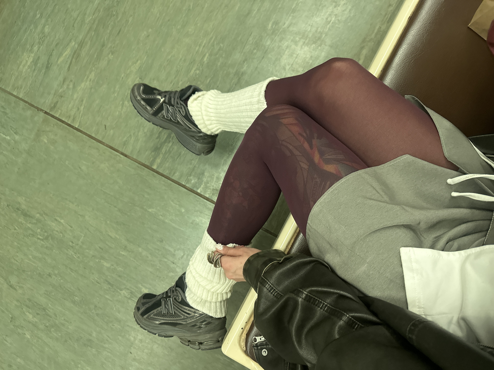 The image shows a person sitting with their legs crossed. They are wearing dark leggings with a subtle pattern, white leg warmers, and dark athletic shoes. The person is also wearing a grey jacket over a white top and a black leather jacket. They are sitting on a brown seat, possibly in a public transportation vehicle or waiting area. The floor is a light green tiled surface. A hand with several rings is visible, resting on one of the leg warmers. The lighting suggests it might be indoors, possibly during the day. There is no visible text in the image.