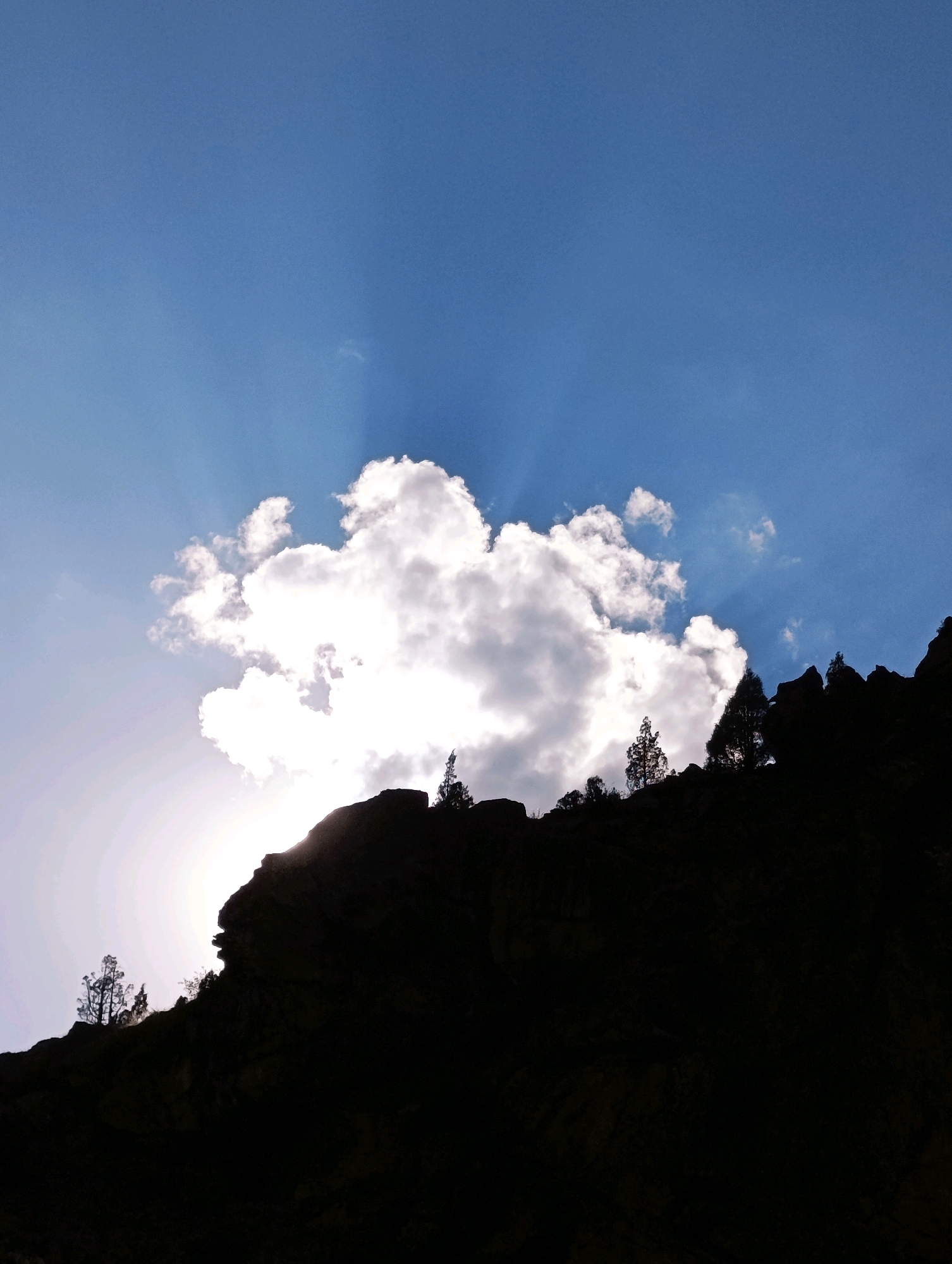 A low-angle, full shot captures a dramatic scene in Stingri, India, where a large, bright cloud partially obscures the sun, casting rays of light across a deep blue sky. The foreground is dominated by the silhouetted dark mass of a rocky hillside, jagged and uneven, with sparse evergreen trees dotting its upper edge. The sun's brilliant glare emanates from behind the cloud, creating a strong contrast with the darkened landscape.