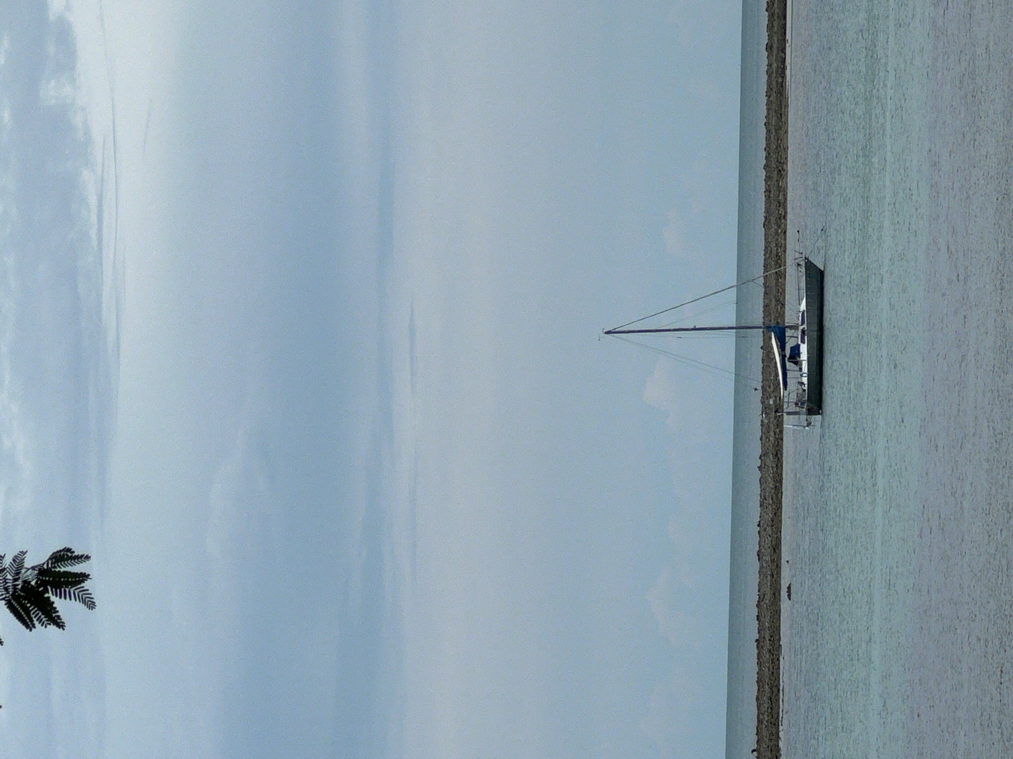 A sailboat is anchored in calm, turquoise waters off the coast of Baan Hua Thanon, Thailand. The sky is overcast with light gray clouds. In the foreground, the water is a muted blue-green. A low, rocky breakwater runs horizontally across the middle ground, separating the calmer inshore waters from the slightly rougher offshore sea. The sailboat, a monohull with a white hull and a blue bimini top, is positioned to the right of center in the frame. Its mast is tall and slender. A few dark green leaves from a tree branch hang from the top left corner of the image. The overall impression is one of stillness and quiet, with a sense of vastness in the open sky and sea.