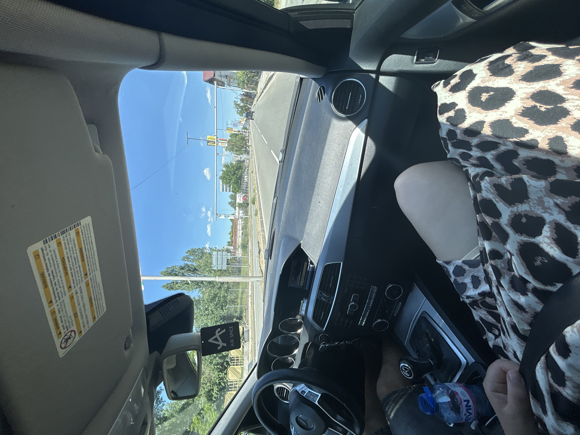 The image shows the interior of a car, specifically the driver's side. The car is stopped at a traffic light in Yahinovo, Bulgaria. A woman is in the passenger seat wearing a leopard-print dress and a seat belt. A bottle of water is visible between the driver and passenger. The dashboard, steering wheel, and other interior components are also visible. Exterior elements seen through the windshield include traffic lights, street signs, buildings, and trees. The weather appears sunny.