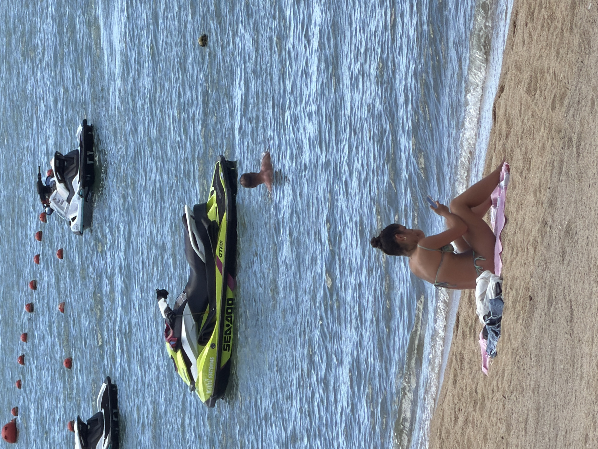 The image depicts a sunny day at a beach in Baan Bo Phut, Thailand. The scene is set along the shoreline where the calm, blue water meets the sandy beach.
Several jet skis are visible in the water, suggesting recreational activities. Two jet skis are closer to the shore, with one appearing to be a yellow and black Sea-Doo GTI model, identifiable by the text "SEA-DOO" on its side. Another jet ski, white with black accents, is further out. In the distance, a few red buoys are lined up in the water.
On the sandy beach, a woman is seated, facing the water. She is wearing a bikini and appears to be holding a mobile phone. Her legs are crossed as she sits on a pink towel, with some clothes, including a pair of jeans and a white top, placed beside her. Her hair is tied up in a bun. The lighting suggests it is daytime, likely midday given the bright conditions and lack of distinct shadows. The overall atmosphere seems relaxed, indicative of a vacation or leisure outing at the beach.