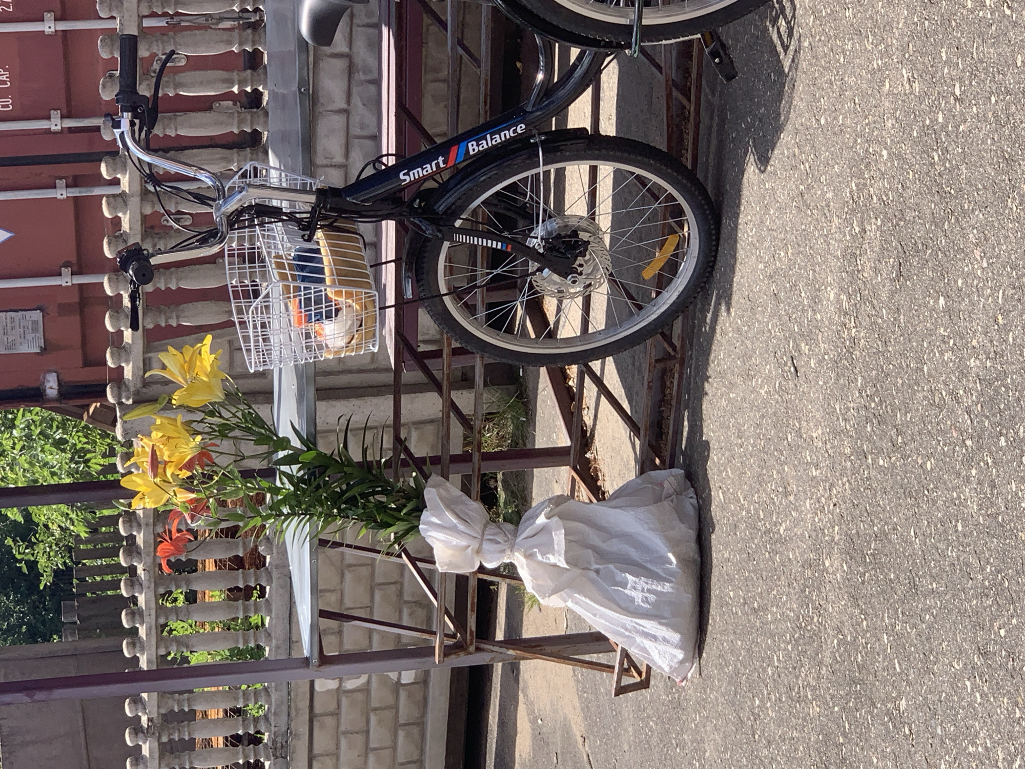 A black bicycle with a white wire basket is parked on an asphalt surface next to a metal structure. The basket contains several items, including what appears to be a yellow toy and some rolled-up materials. A bouquet of lilies, with yellow and orange flowers, is tied with white fabric and secured to the metal structure. The scene is outdoors, with a concrete fence and greenery visible in the background. A large red shipping container is also partially visible. The photograph was taken in Kapyĺ, Belarus.