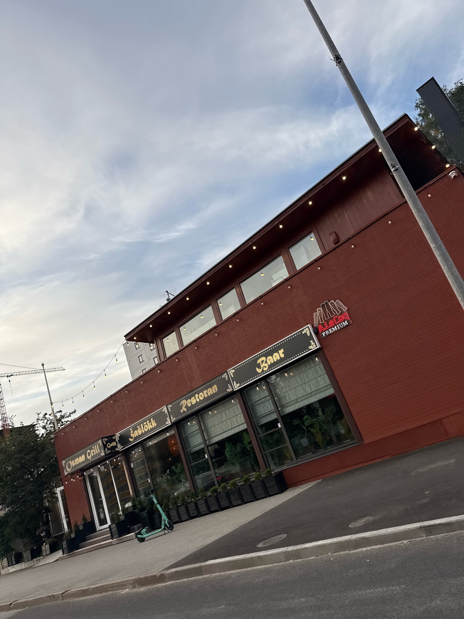 The image shows a brick building with a red facade, tilted upwards to show the sky. The building has large windows and is adorned with string lights. A sign on the building reads "Osman Grill", "Sašlõkk", "Baar", and "Restoran". Another sign shows "A.LeCoq PREMIUM". In front of the building, there is a sidewalk and a street. A green electric scooter is parked on the sidewalk. A construction crane is visible in the background on the left. The sky is cloudy and a utility pole is prominent in the foreground on the right. The scene appears to be in an urban environment in Tallinn, Estonia, likely a commercial area with restaurants and bars.