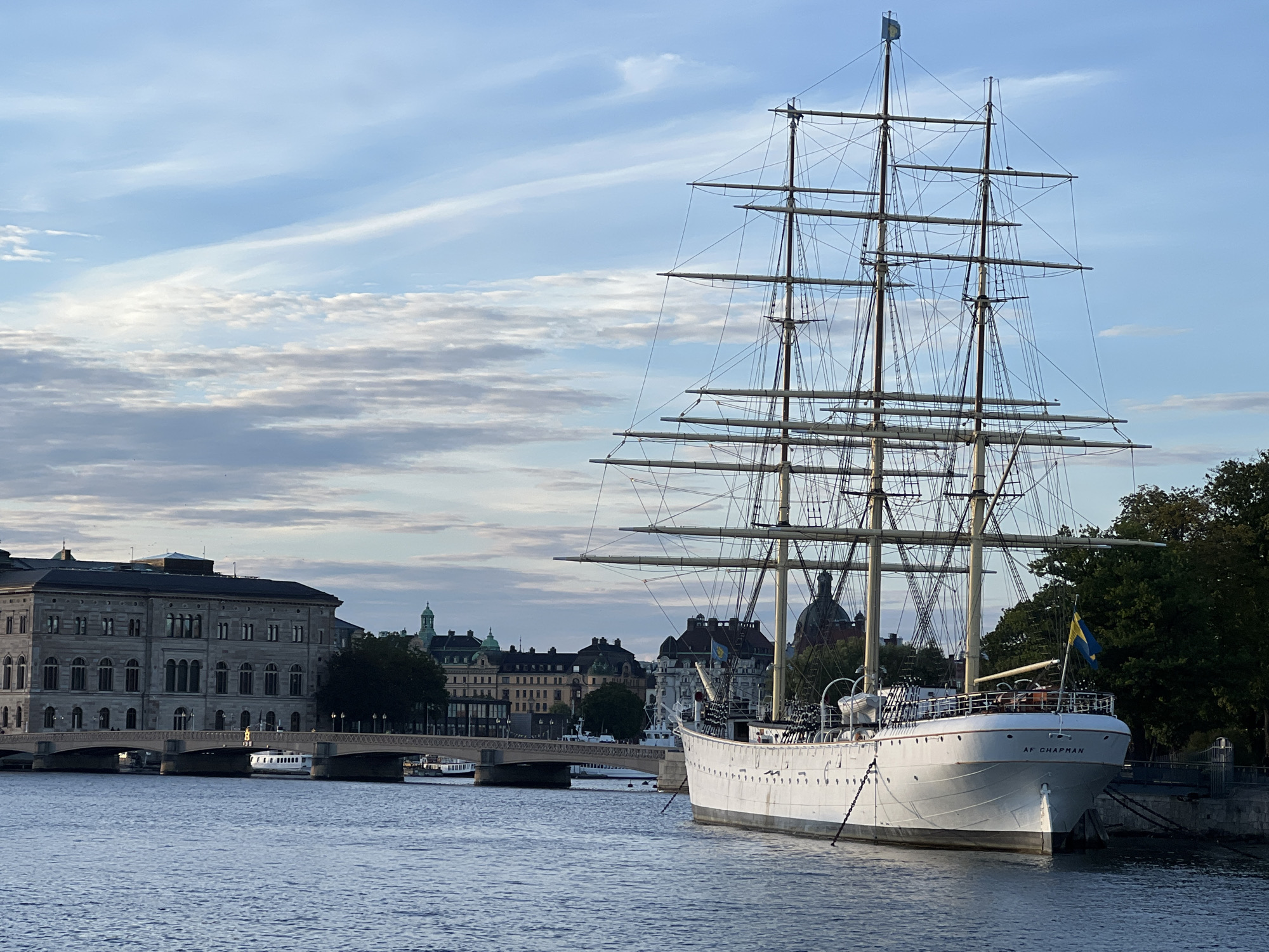 The image shows the Af Chapman sailing ship moored in Stockholm, Sweden. The ship is a large, white, three-masted vessel with numerous sails and rigging visible. It is positioned in a body of water, likely a harbor or canal, with a cityscape in the background. The cityscape includes several buildings of varying architectural styles, a bridge, and some trees. The sky is partly cloudy with a mix of blue sky and white clouds. The overall scene suggests a bright day, possibly late afternoon or early morning given the lighting. The name "AF CHAPMAN" is visible on the bow of the ship.