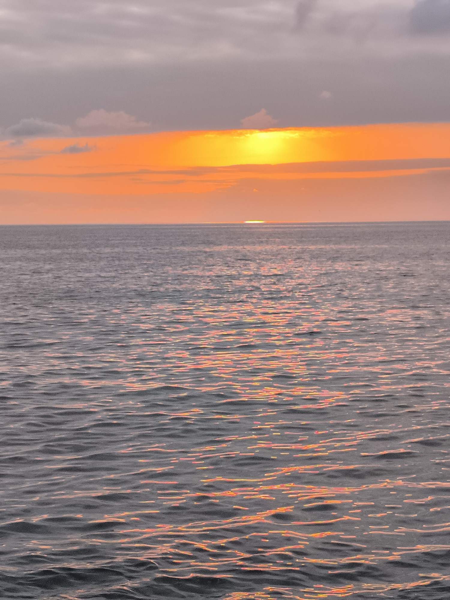 The image depicts a seascape at dusk, with the sun partially visible on the horizon. The sky is filled with clouds, ranging in color from soft gray to warm orange and yellow, indicating the sunset. A bright band of orange light stretches across the sky, with the sun appearing as a small, intense yellow glow just above the water's surface. The sea is calm, with gentle ripples reflecting the colors of the sunset, creating streaks of orange and pink light on the dark water. The horizon line is distinct, separating the sky from the sea. No people, structures, or specific city landmarks are visible.