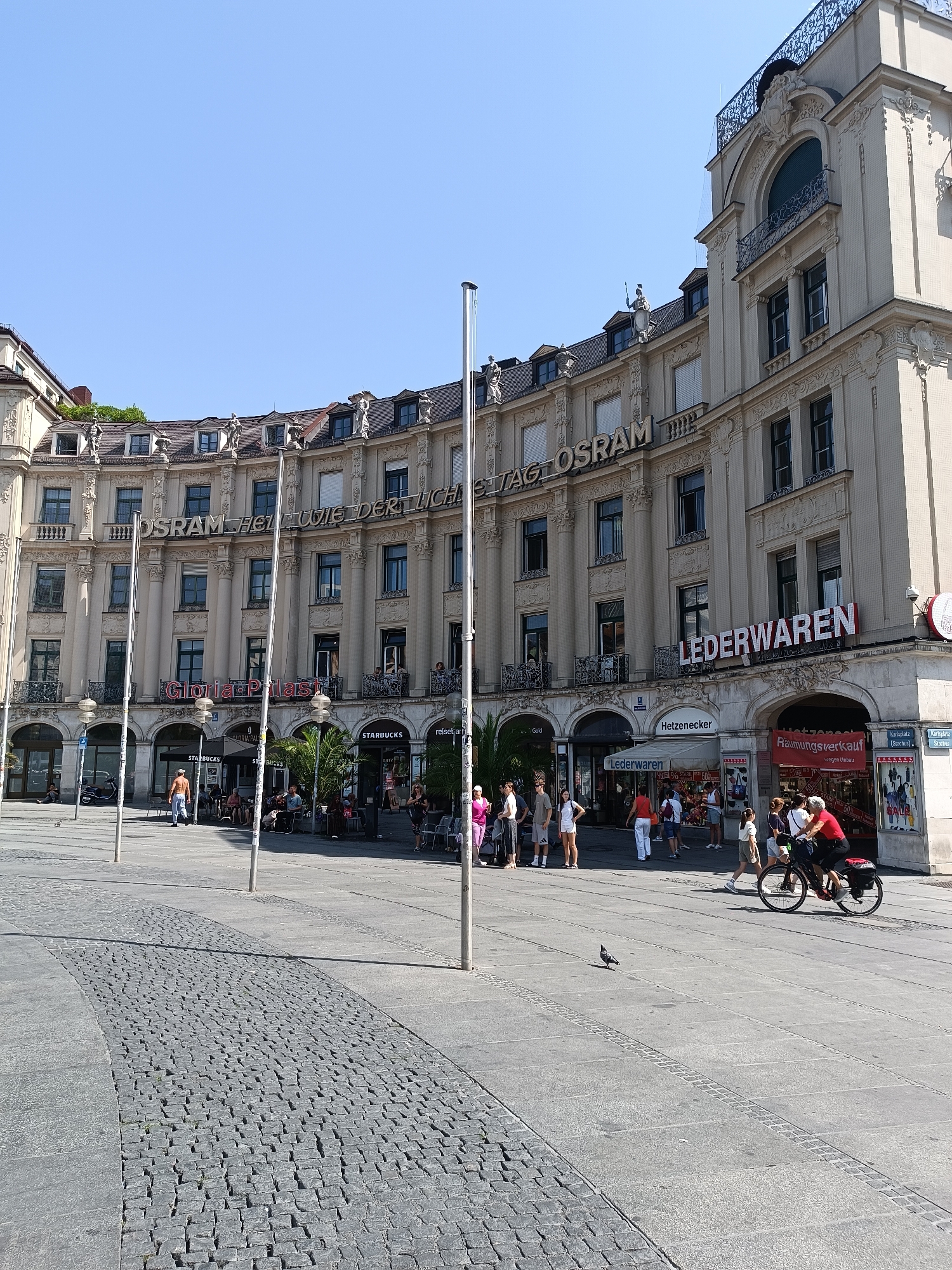 The image shows a street scene in Munich, Germany, specifically on Kaufingerstraße, a prominent shopping street. The iconic Osram building, with its ornate facade and the inscription "OSRAM HELL UND WEISS DER LICHTERSTADT OSRAM", dominates the background. The building features multiple stories, arched windows, and decorative sculptures. In front of the building, there are several shops, including "Lederwaren" (leather goods) and a Starbucks. People are seen walking, sitting at outdoor tables, and cycling on the cobblestone street and pavement. The sky is clear and blue, indicating a sunny day. The overall scene depicts a busy urban environment with a mix of historical architecture and modern commercial activity.