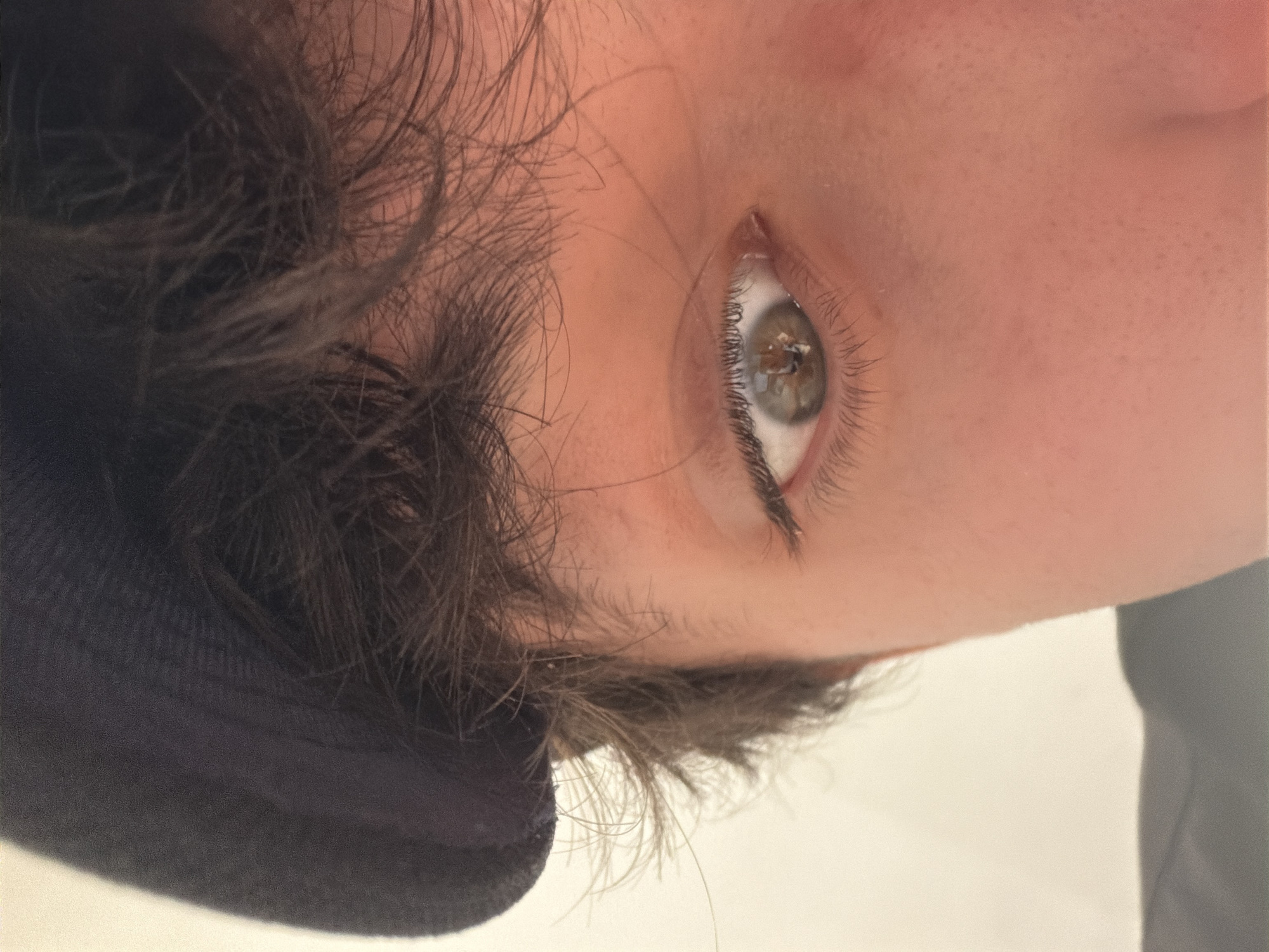 The image is a close-up of a person's eye and forehead, wearing a black beret. The lighting suggests an indoor setting with a light-colored background. There are no discernible elements that would indicate a location in Cancún, Mexico. The subject's skin tone is light, and their hair is dark brown and somewhat messy. The eye is a light brown or hazel color, and the iris shows a reflection of what appears to be a bright light source.