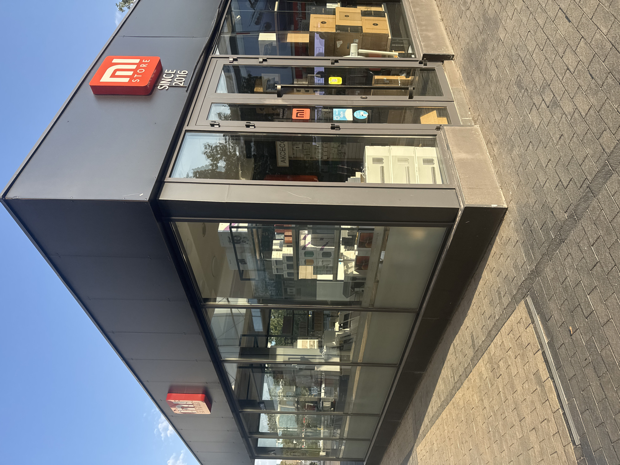 A modern Mi Store storefront is captured in this image, located in Bishkek, Kyrgyzstan. The building features a dark gray facade with large glass windows and double glass doors, allowing a view of the store's interior. A prominent red Mi Store logo is displayed on the upper left of the building, with the text "SINCE 2016" below it. Inside, shelves are stocked with various electronic products and accessories. The exterior is enhanced by a paved walkway made of light brown and gray brick-like tiles, leading up to the entrance. The scene is bathed in the warm light of a sunny day, casting distinct shadows on the ground. The sky above is a clear, bright blue.