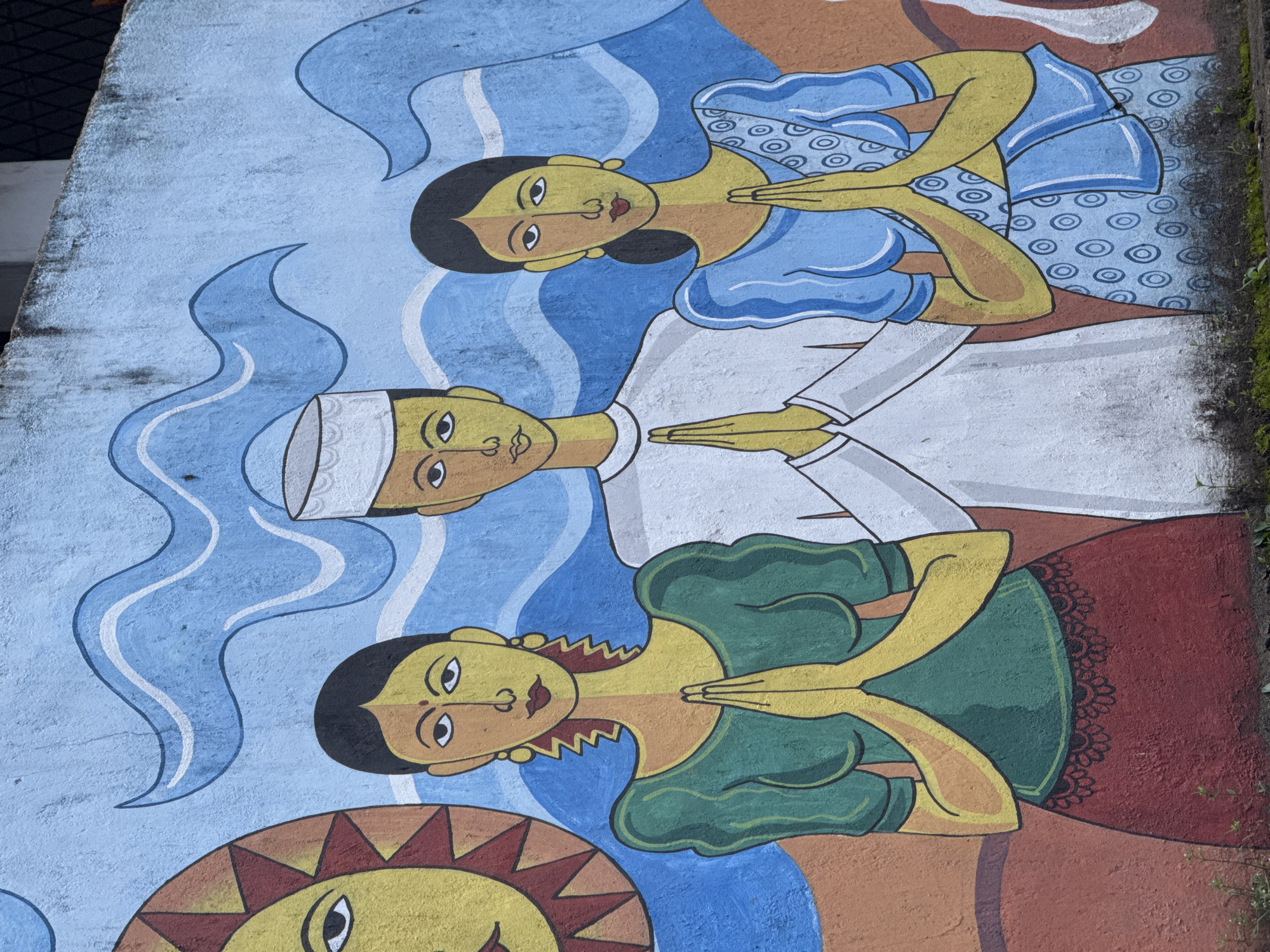 A mural on a rough-textured, light blue concrete wall depicts three figures with yellow faces and dark hair, stylized with simple geometric shapes. The figures are positioned centrally and are all depicted with hands pressed together in a gesture of greeting or prayer. To the left, a female figure wears a green blouse with puffed sleeves and a dark red skirt. Her earrings are yellow and jagged, and her hair is dark and styled in waves. A large, stylized yellow sun with red triangular rays is positioned behind her left shoulder. In the center, a male figure wears a white robe and a white cap with a textured brim. To the right, a female figure wears a bright blue saree with a pattern of concentric circles. Both female figures have a red dot on their forehead. The background behind the figures features stylized blue waves. The mural is located in Dondra, Sri Lanka, a coastal town in the Southern Province known for its lighthouse and ancient Dondra Temple.