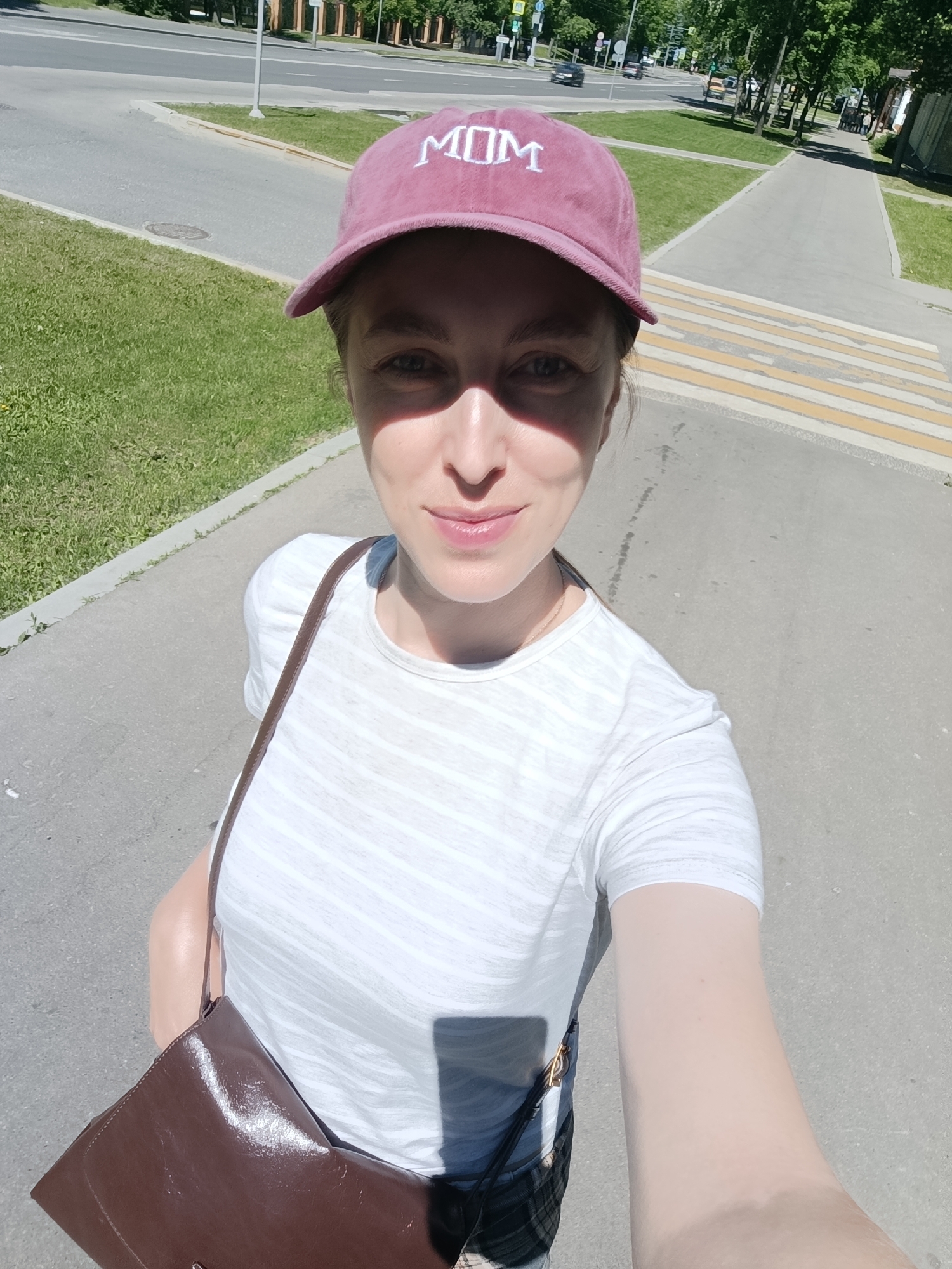 A fair-skinned woman is depicted in a brightly lit mid-shot selfie, standing on a paved sidewalk under strong sunlight in Moscow, Russia. She wears a maroon baseball cap with "MOM" embroidered in white capital letters on the front, casting a shadow over her upper face. Her lower face, including her mouth which holds a subtle smile, is illuminated. She is dressed in a white t-shirt with subtle horizontal heathered stripes and wears a glossy, dark brown shoulder bag with a thin strap across her body. A thin necklace chain is visible around her neck.
The background reveals a typical urban environment on a clear day. To the woman's left, a narrow strip of green grass separates the sidewalk from an asphalt road. The road features white lane markings and, in the distance, shows several moving vehicles, including a black car and an orange/yellow vehicle, along with visible traffic lights and streetlights. To her right, the paved sidewalk continues towards a distinctive white and yellow striped pedestrian crossing (zebra crossing). Beyond the sidewalk and road, a broader green area with numerous mature trees lines the street, partially obscuring buildings in the far background. A grey utility pole is visible near the road's edge. The strong sunlight creates clear shadows throughout the scene. The white and yellow pedestrian crossing stripes are characteristic of road markings found in Russia, confirming the location as Moscow.