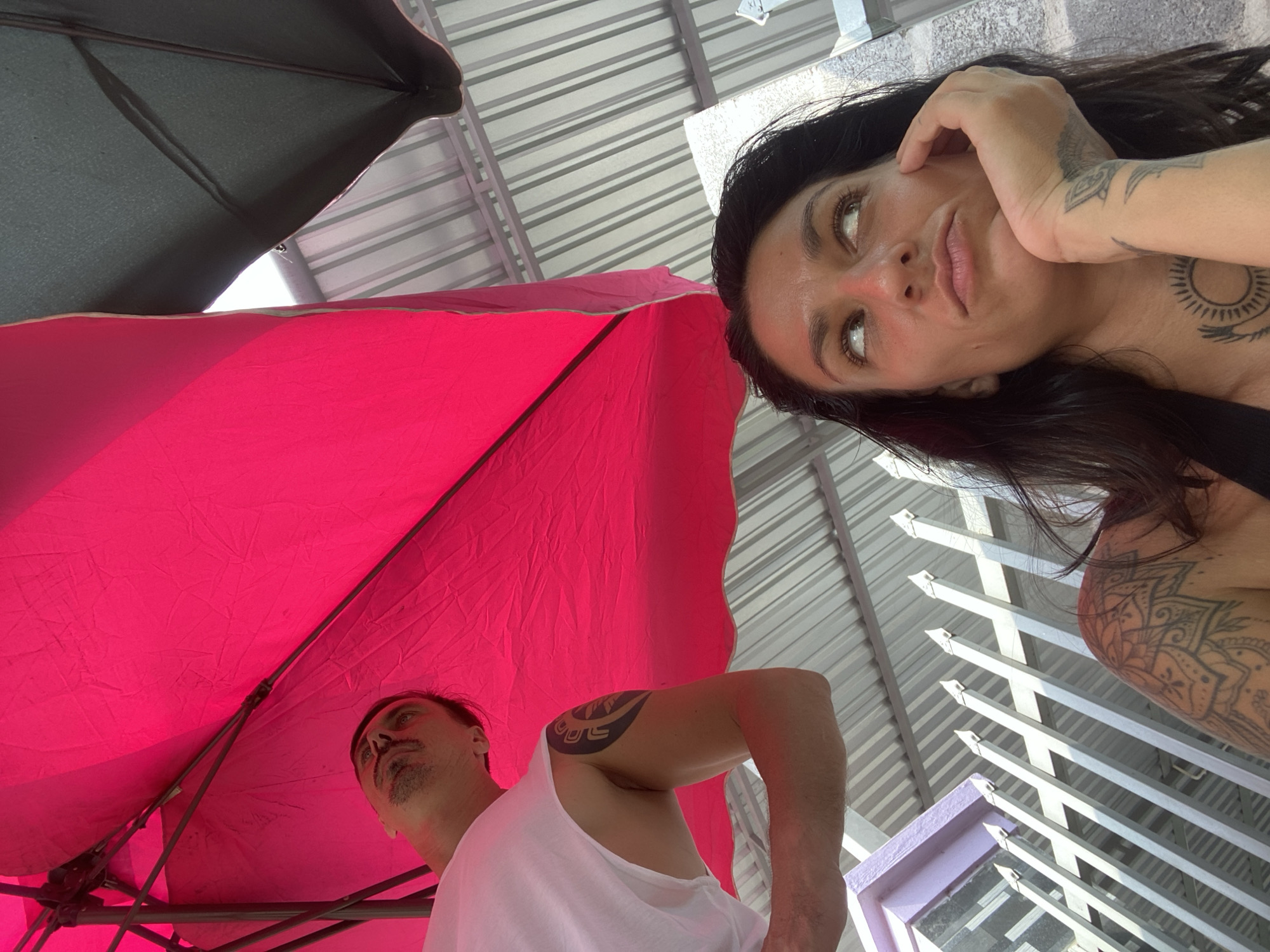 This is a ground-level, outdoor, medium shot of two people under a pink umbrella, possibly at a cafe or restaurant in Mũi Né, Vietnam. In the foreground, a woman with dark hair is looking up and to her right with a pensive expression, her left hand resting on her chin. She has tattoos on her left shoulder and chest, and a tattoo on her right forearm. Behind her and to the left, a man wearing a white tank top is also looking up. He has a goatee and a tattoo on his left arm. The background consists of a corrugated metal roof structure and a white metal fence. The lighting suggests it is daytime.