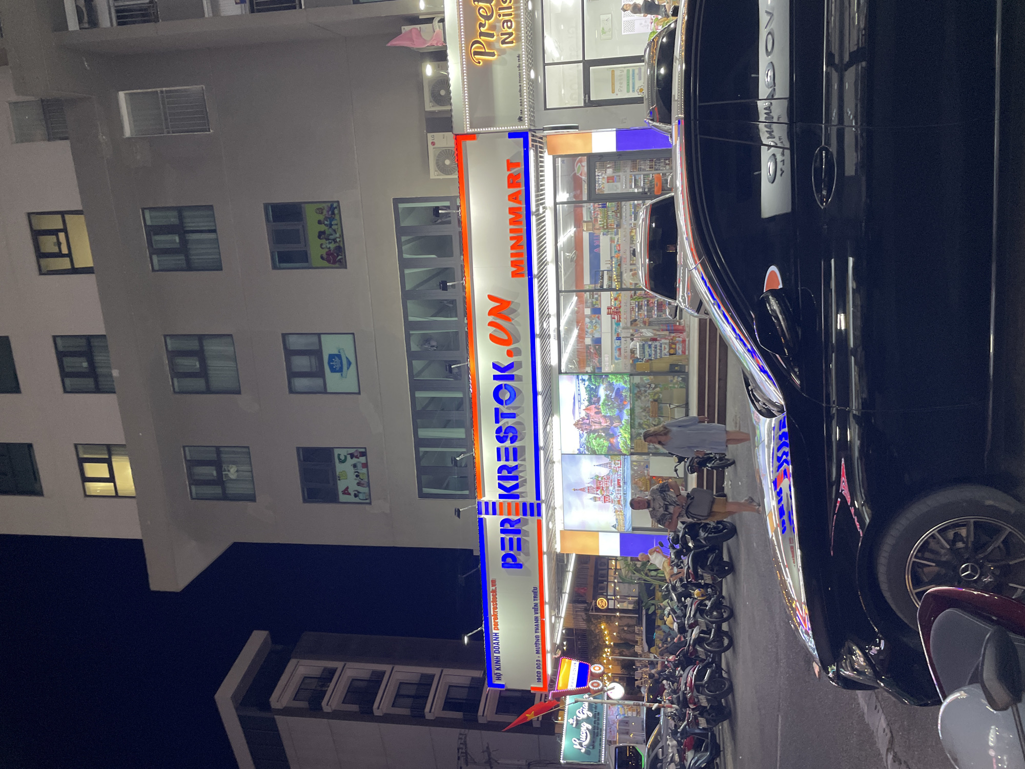 This image captures a nighttime street scene in Nha Trang, Vietnam. The focus is on the facade of a "PEREKRESTOK.VN MINIMART" store, illuminated by bright neon lights. The store's signage is prominent, featuring red, white, and blue lettering. In front of the minimart, there is a display of various products on shelves visible through the glass storefront. Two individuals, a man and a woman, are walking in front of the minimart. To the left of the minimart, another shop with the signage "Luong Gia" is visible, also lit up. A line of parked motorcycles is present on the left side of the street, and a black car is parked in the foreground, partially obscuring the view of the minimart. The building above the minimart has several floors with windows, some of which are illuminated. The overall environment suggests a bustling urban area at night.